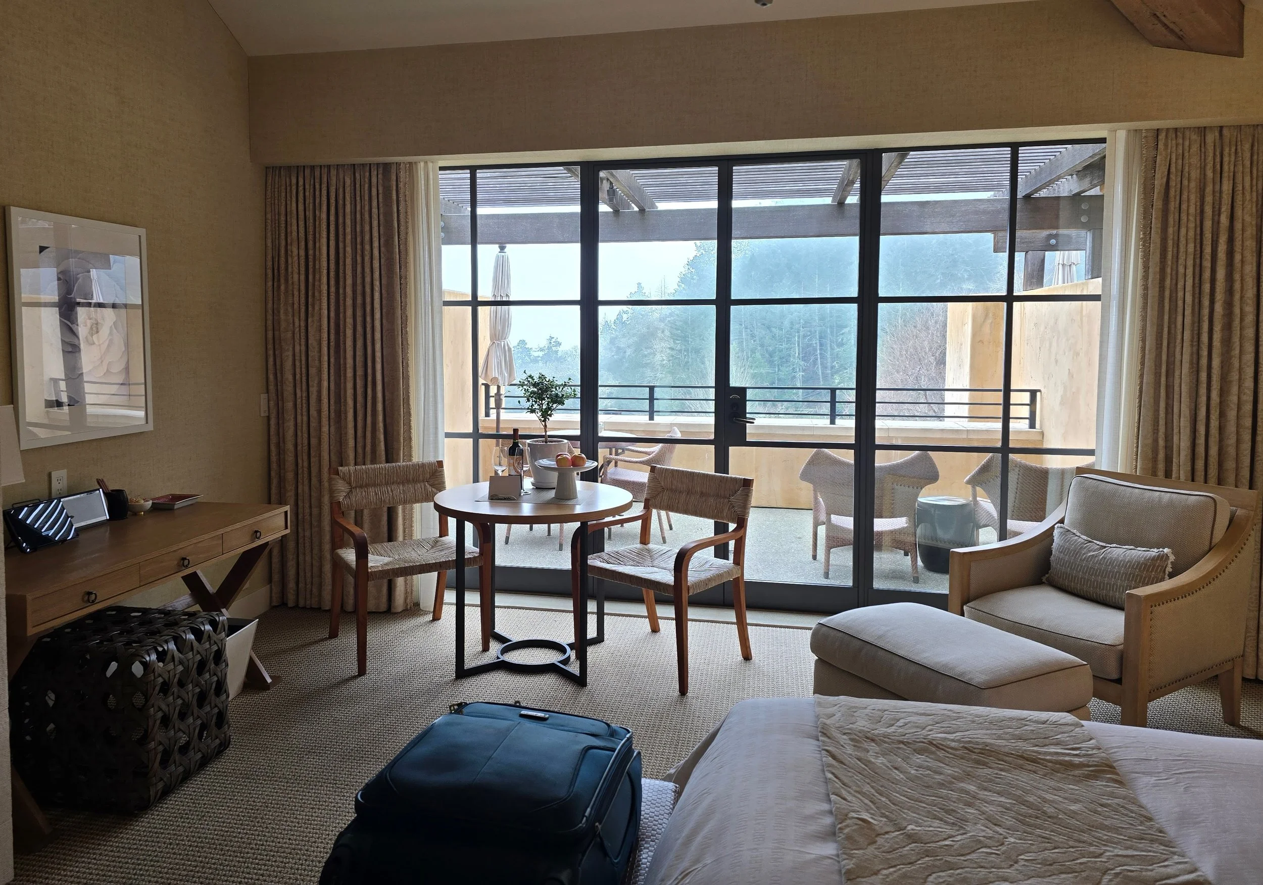 Hotel room with sliding glass door leading to balcony with outdoor chairs and umbrellas, a small round dining table with wine and apples, a bed partially visible, a beige armchair with matching ottoman, and a luggage on the floor.