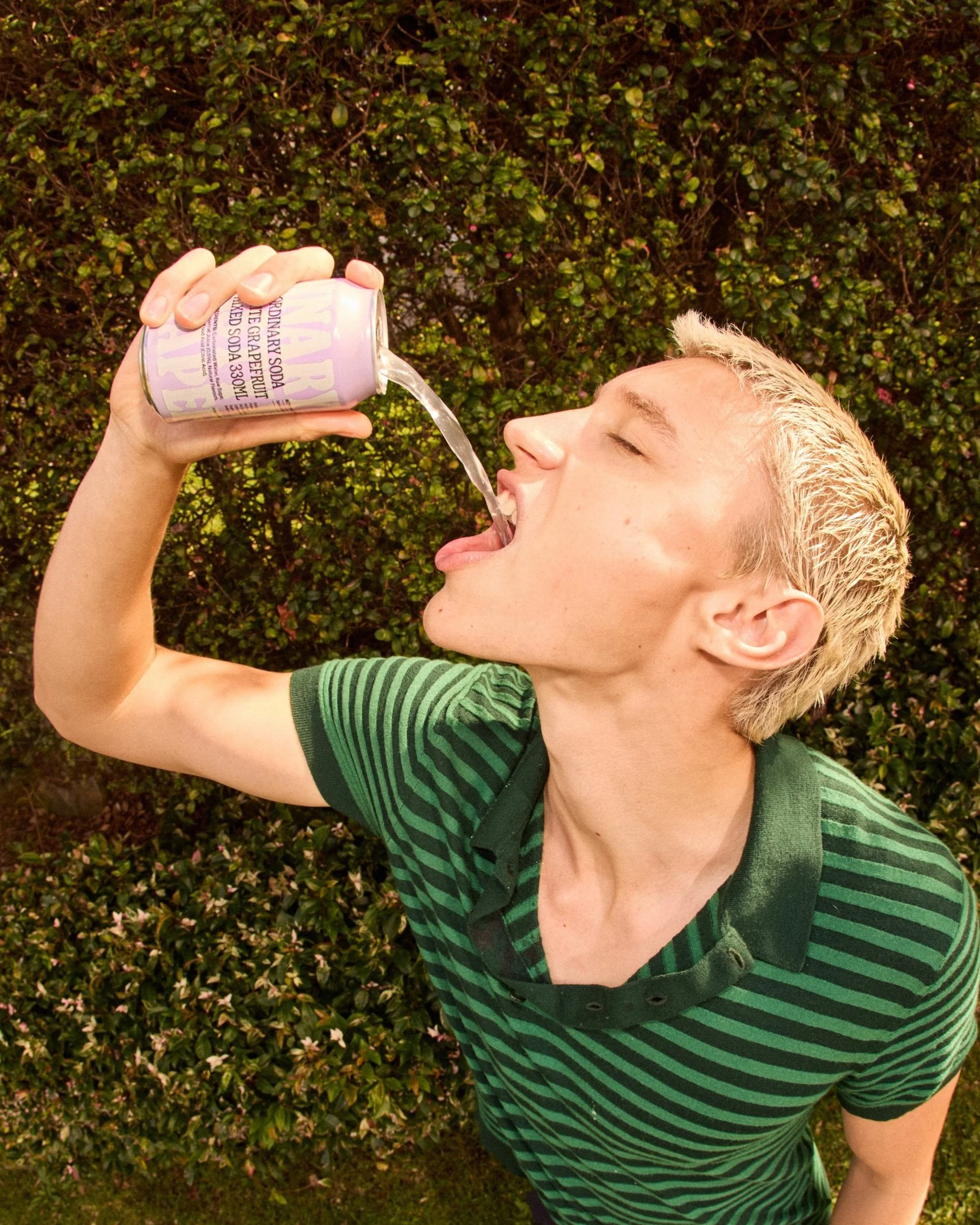 A young person with blond hair wearing a green striped shirt is tilting their head back and pouring sparkling water from a can into their open mouth outdoors with green foliage in the background.