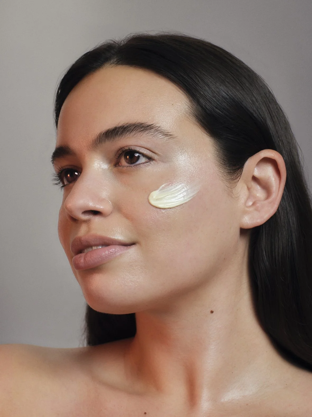 Close-up of a woman with dark hair and natural makeup, applying sunscreen to her cheek.