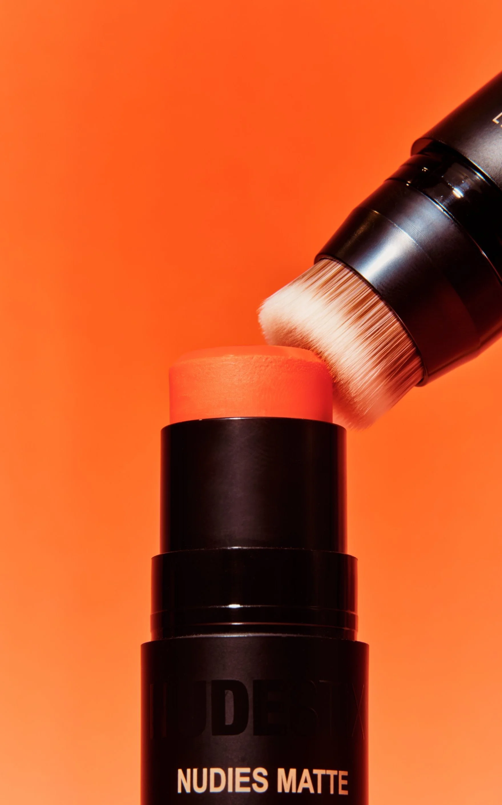 Close-up of a black cylindrical makeup product container labeled 'NUDES MATTE' with a pinkish-orange makeup sponge on top and a makeup brush applying product onto the sponge, against an orange background.