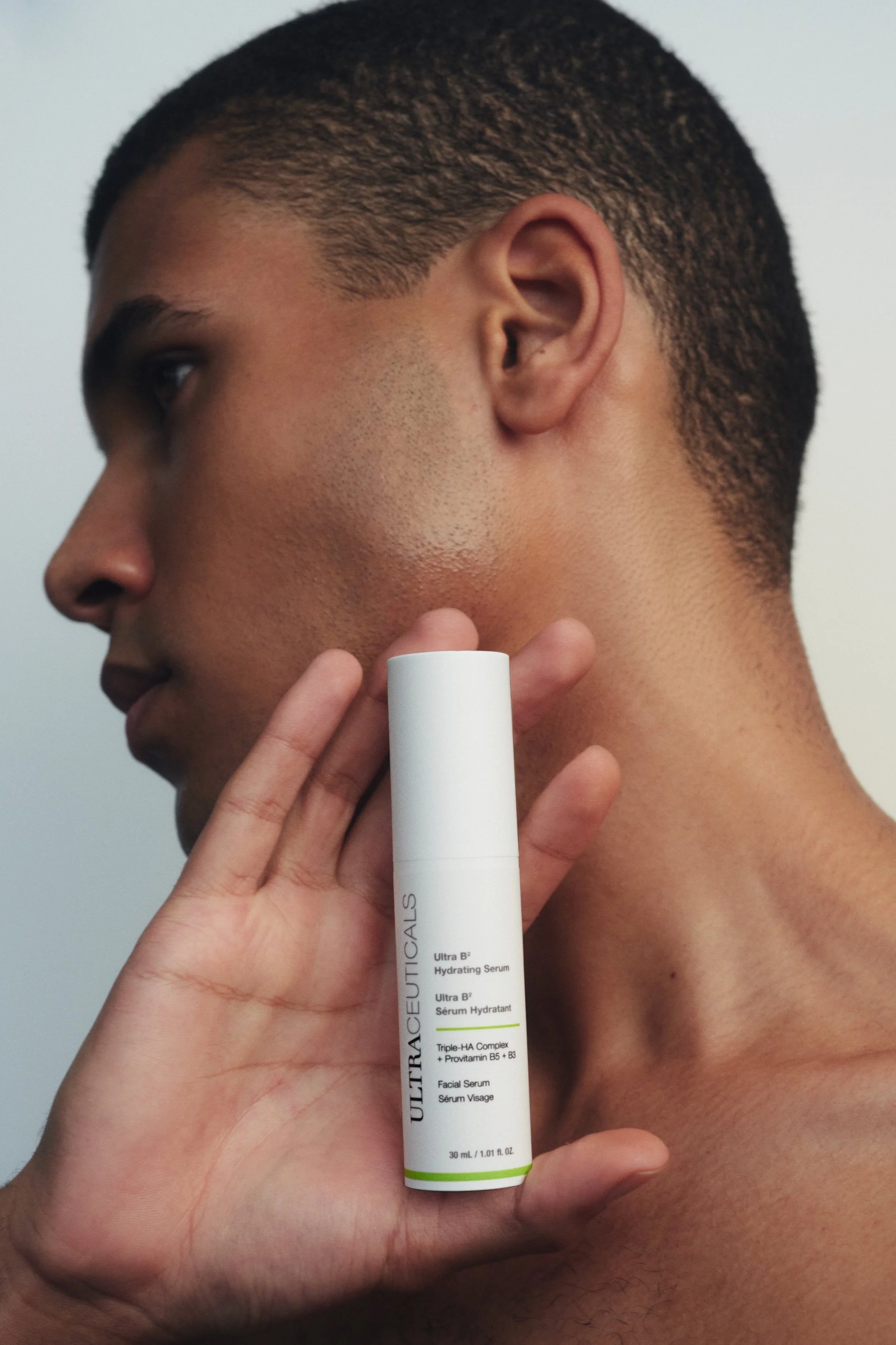 A person holding a white tube of Ultra B3 Hydrating Serum against the side of their face, with their head turned to the left. The serum bottle is labeled with product details and is positioned near the jawline.