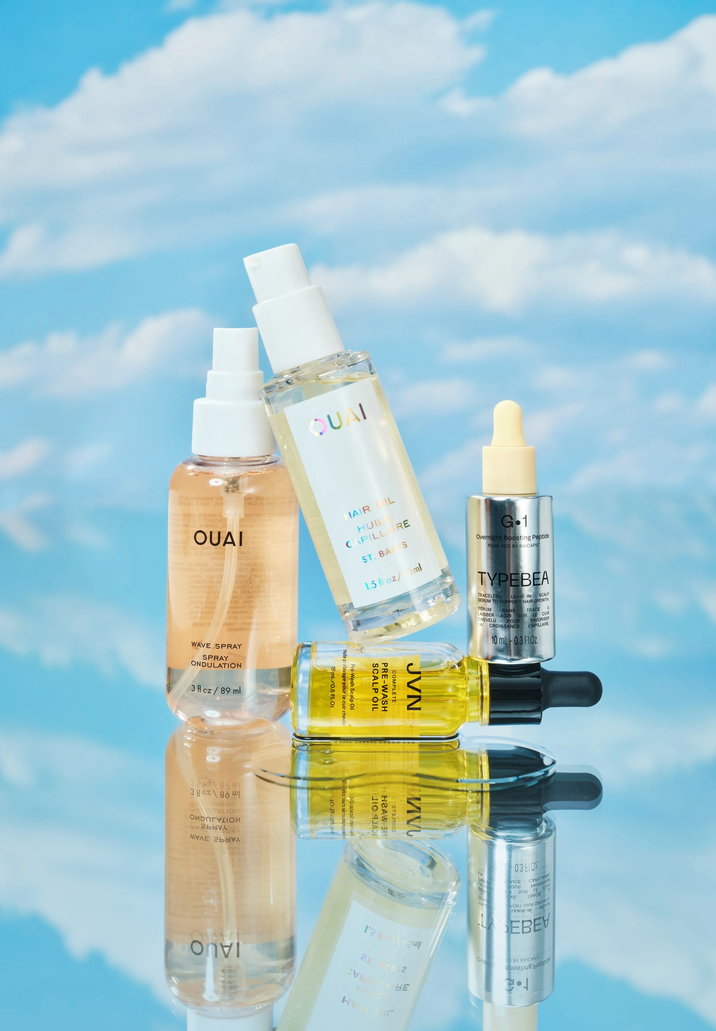Collection of hair care products including Ouai wave spray, Ouai hair oil, Tyeebe scalp oil, and Jyun scalp oil, arranged on a reflective surface with a cloudy sky background.