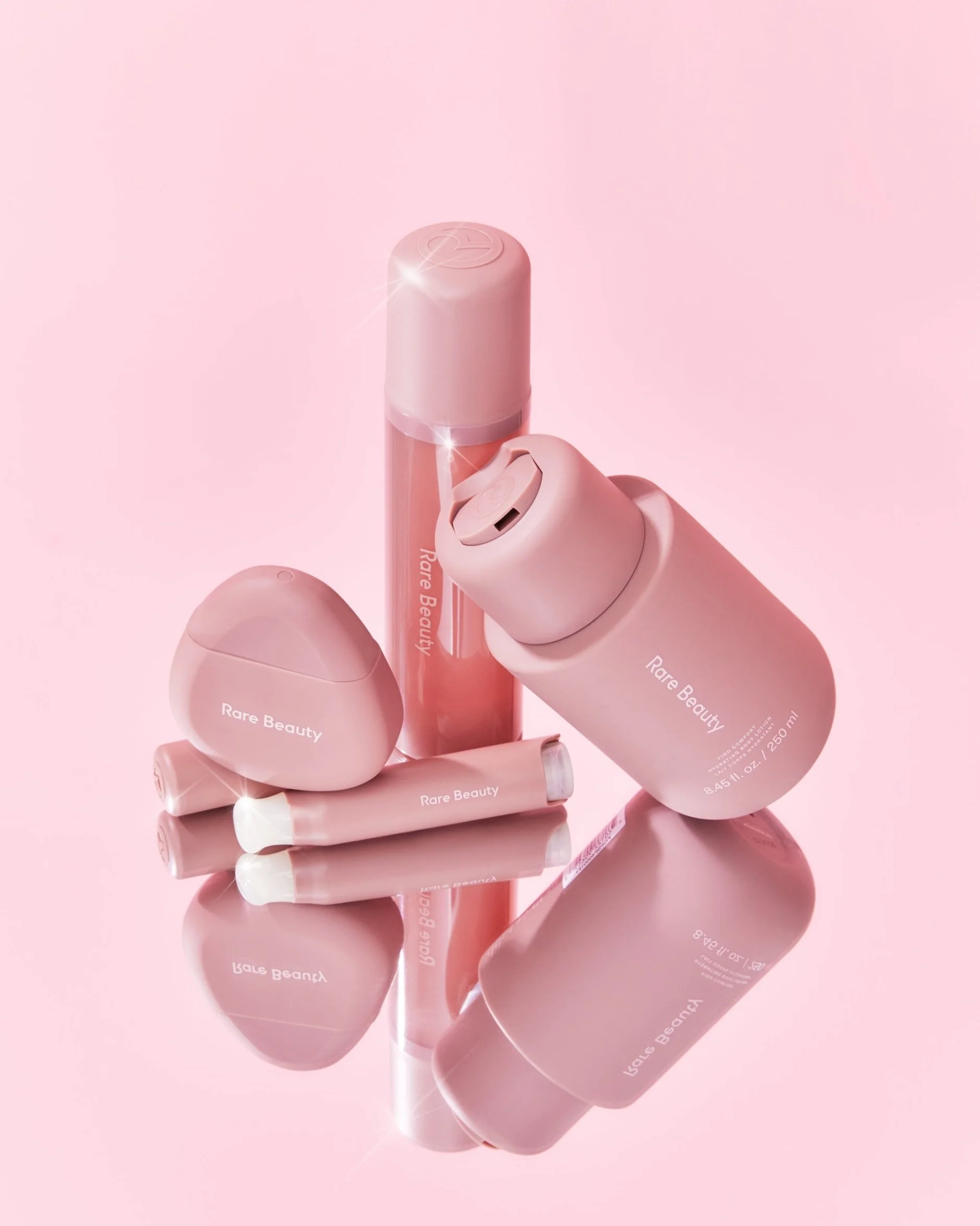 Collection of pink beauty products including a spray bottle, a small container, and two tubes, all labeled 'Rare Beauty' on a pink reflective surface.