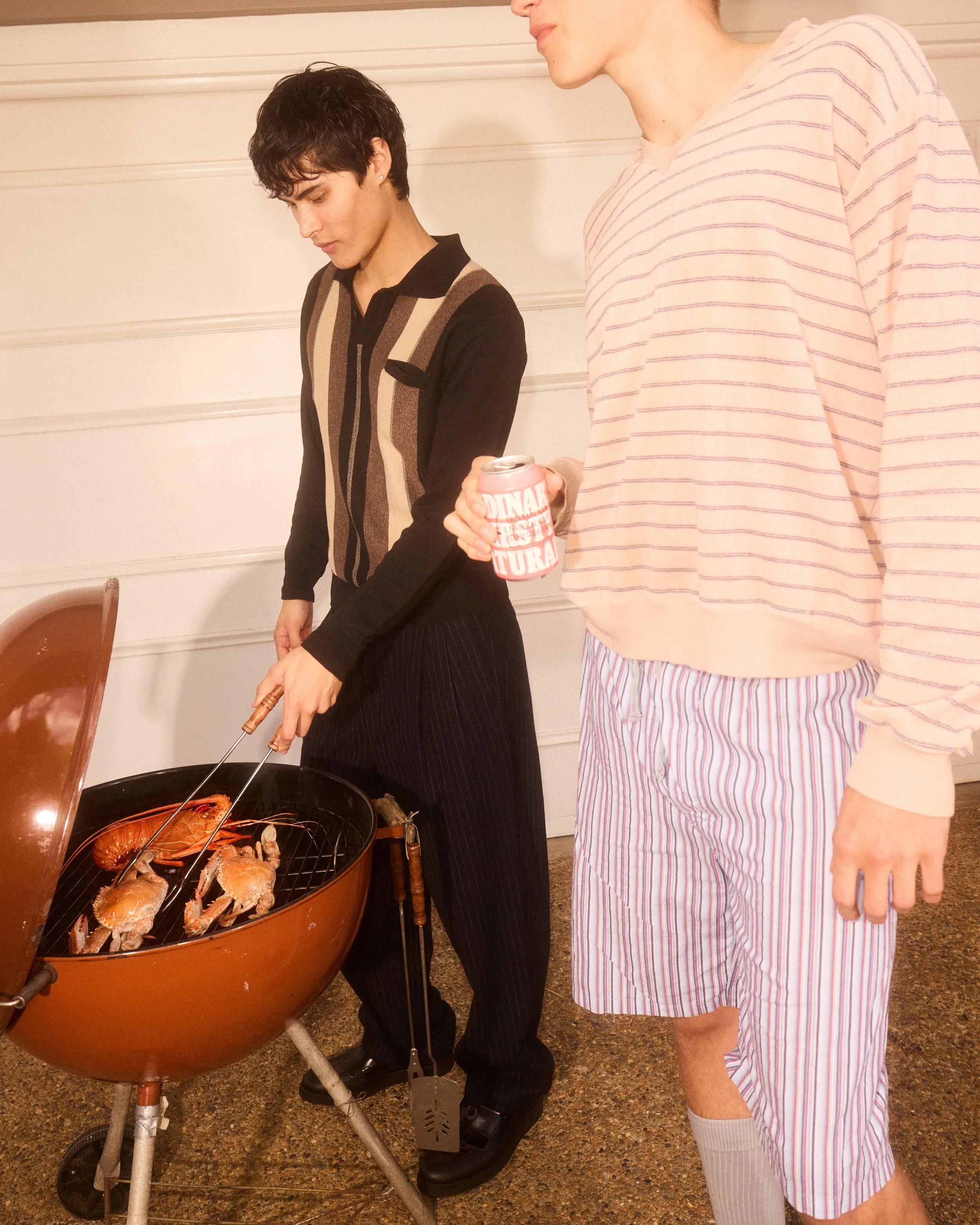Two women standing near a round outdoor grill with seafood. One woman is grilling and holding a pair of tongs, while the other woman is holding a can and wearing pajamas.
