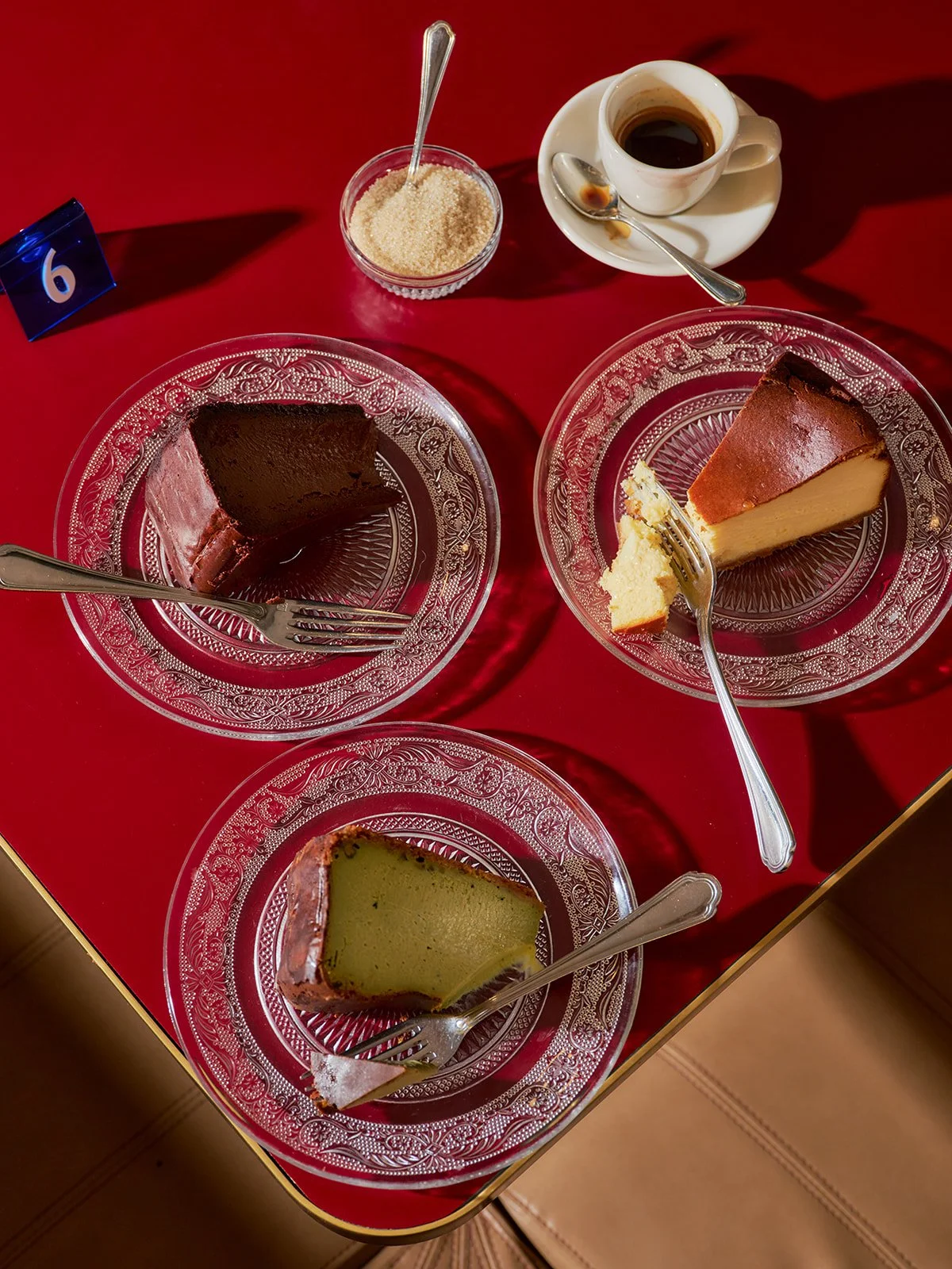 Three plates with different slices of cake, a cup of coffee on a saucer with a spoon, a small bowl of sugar with a spoon, and a blue table number card.
