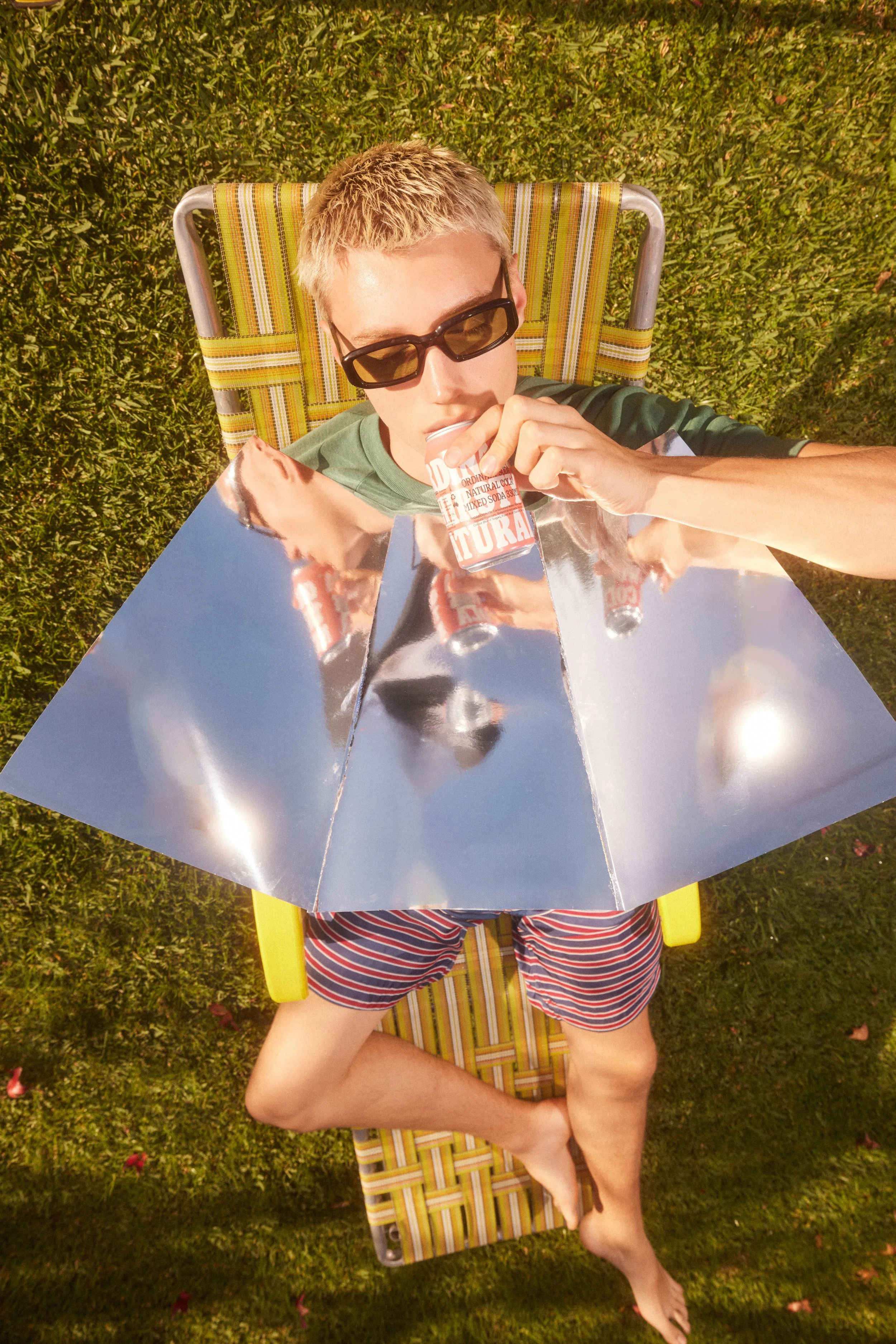 A young man with short blond hair and sunglasses lying on a striped lounge chair outdoors on grass, drinking from a soda can into his mouth, with a reflective square mirror placed on his chest.