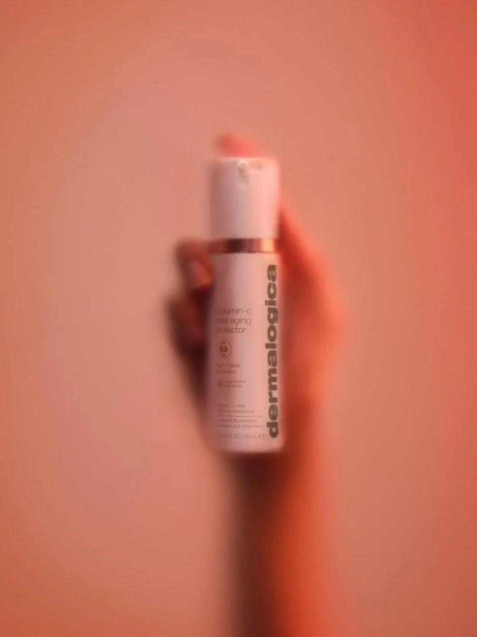 A hand holding a white bottle of DermaE antioxidant-C and collagen booster, with a pink background.