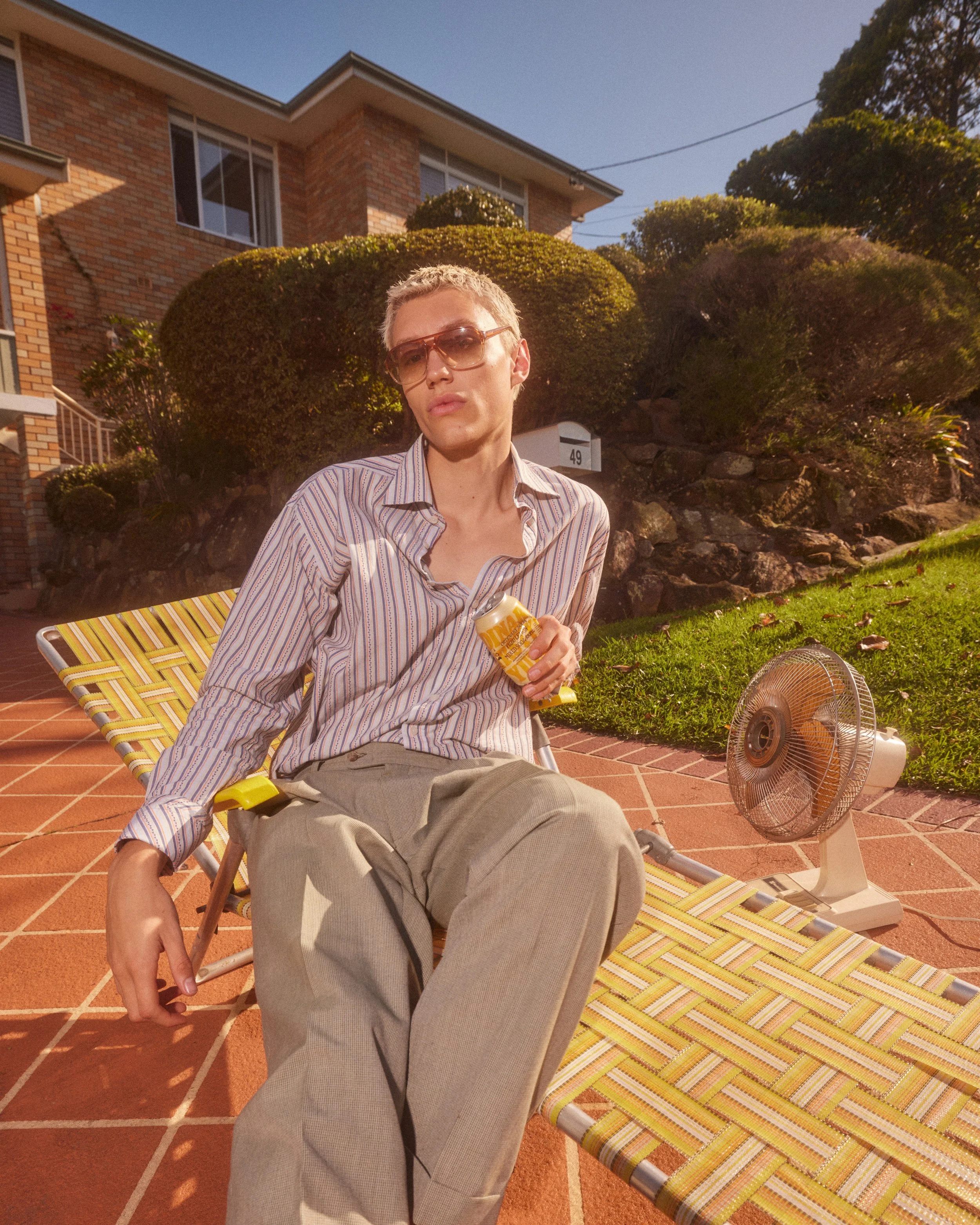 A person with short blonde hair and sunglasses sitting on a yellow-striped lounge chair outside on a brick patio, holding a yellow can, with a small fan nearby and a background of a brick house, plants, and a blue sky.