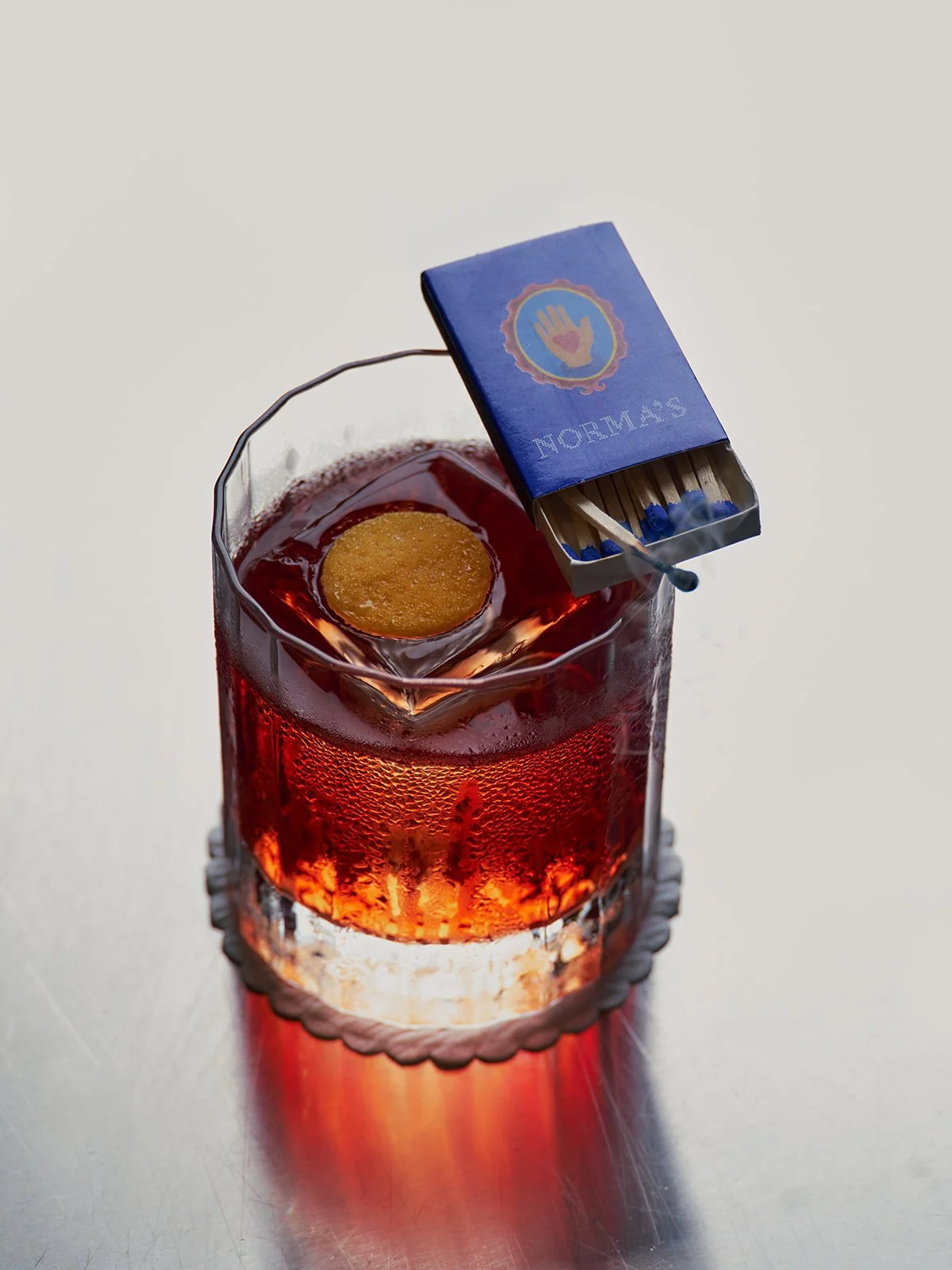 A glass of red beverage with ice and a lemon slice, placed on a clear textured coaster on a metal surface, with a matchbox of matches resting on the ice.
