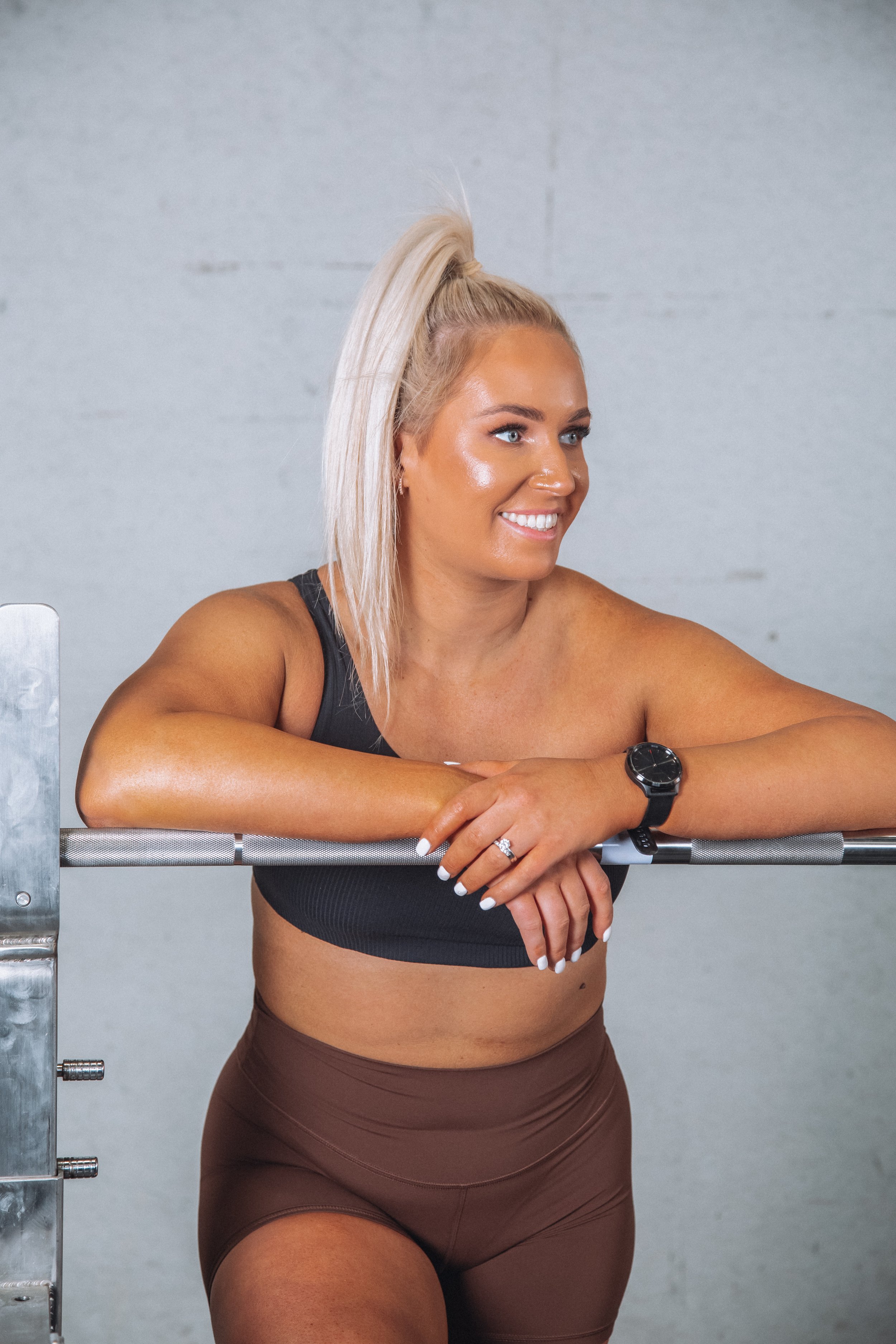 A smiling woman with blonde hair tied in a high ponytail, wearing a black sports bra and brown shorts, leaning on a barbell in a gym.