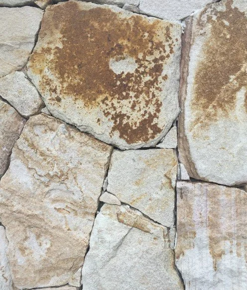 Close-up of a stone wall with irregularly shaped beige and brown stones, some with rust-colored patches and cracks.
