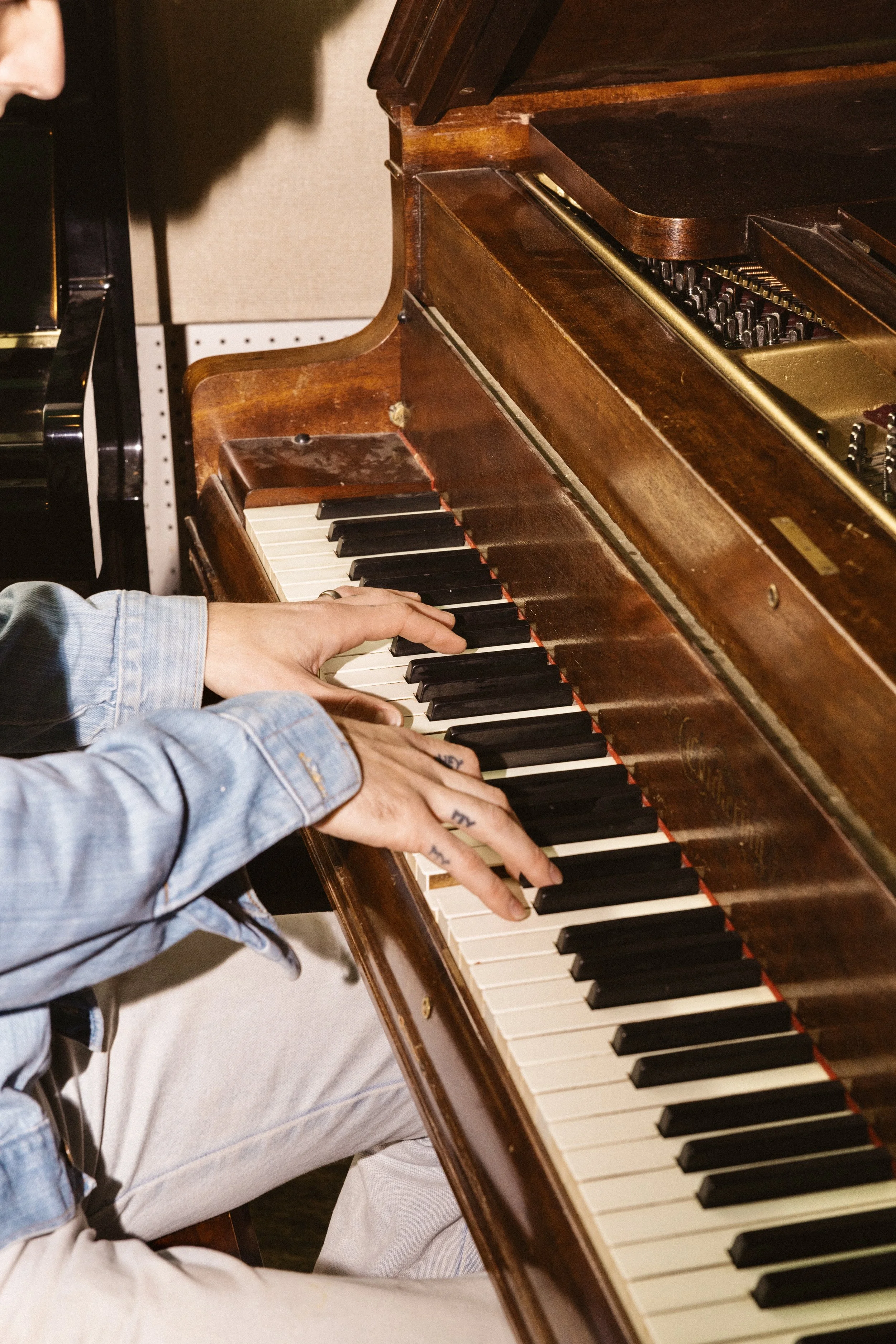 A person playing a wooden upright piano with black and white keys, wearing a denim jacket and beige pants.