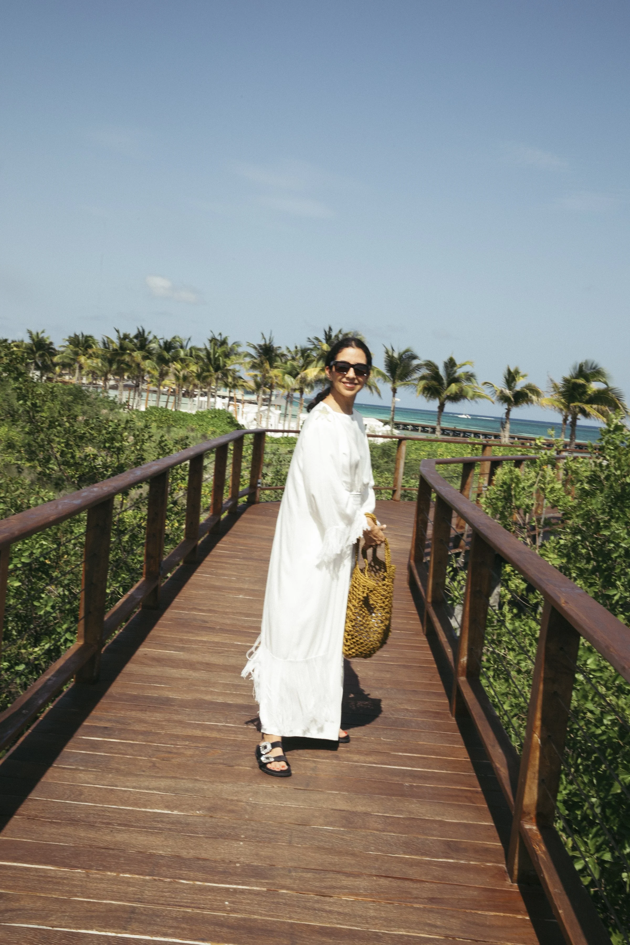A woman in a long white dress and sunglasses standing on a wooden pathway surrounded by greenery with palm trees, overlooking the ocean under a clear blue sky.