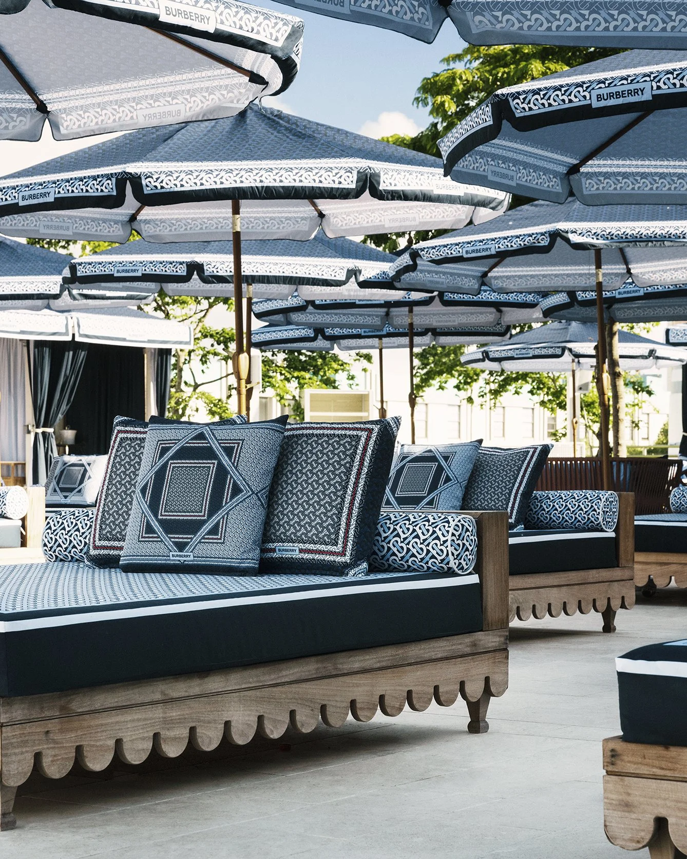Outdoor seating area with wooden sofas and black-and-white patterned cushions and pillows, shaded by large Burberry umbrellas, and surrounded by trees and a clear blue sky.