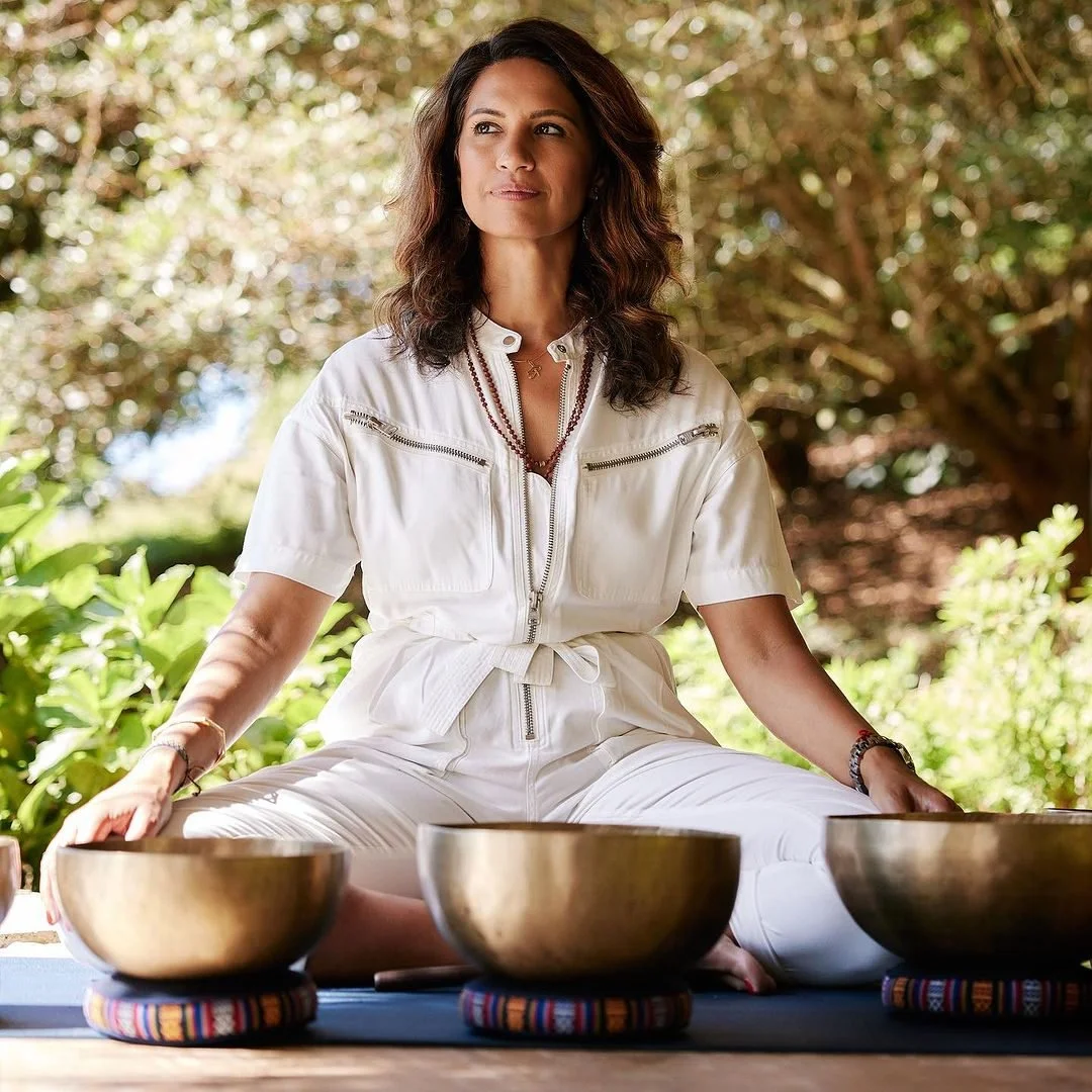 A woman sitting outdoors on a yoga mat, surrounded by greenery, with singing bowls in front of her.
