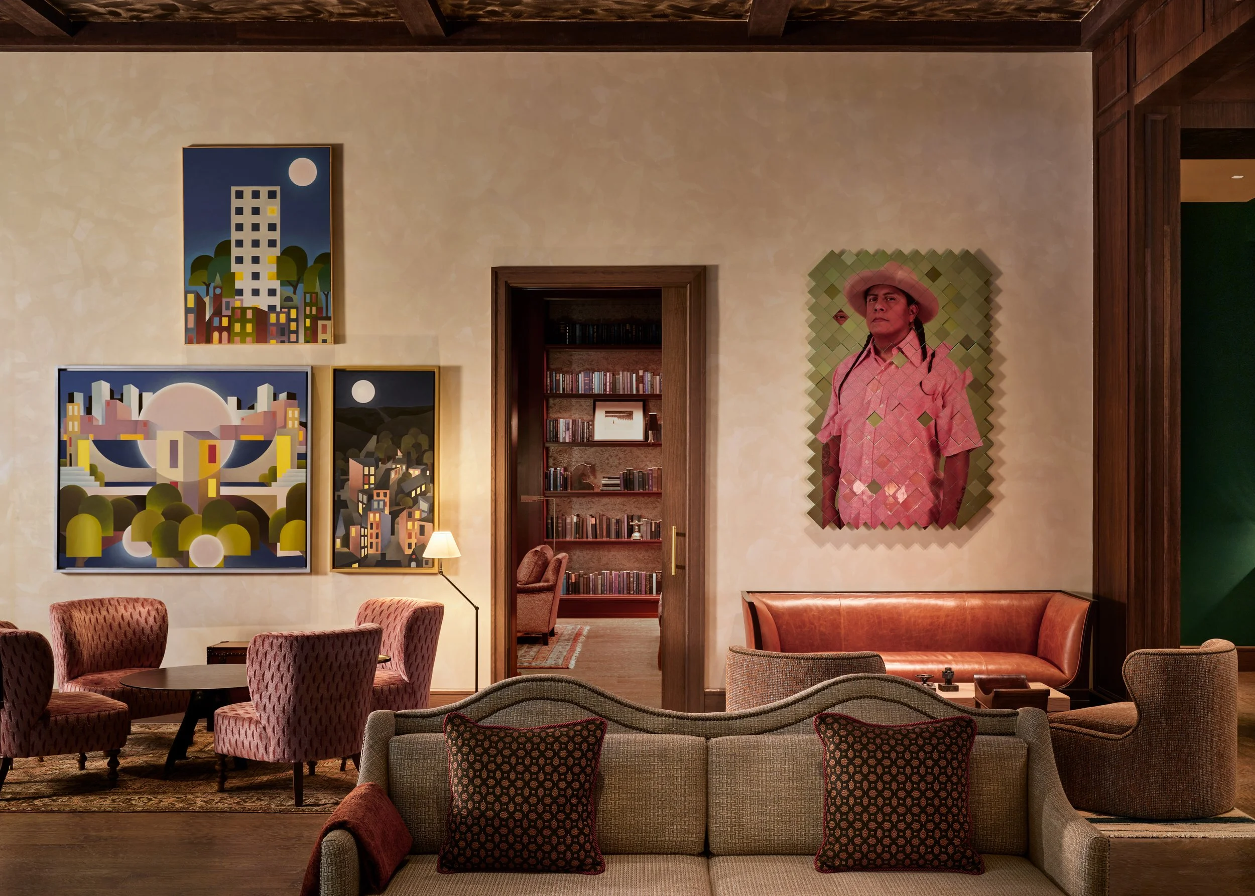 Living room with beige walls, wall art including paintings of buildings and cityscapes, a portrait of a man in a hat and patterned shirt, bookshelves, and furniture like chairs and a sofa.