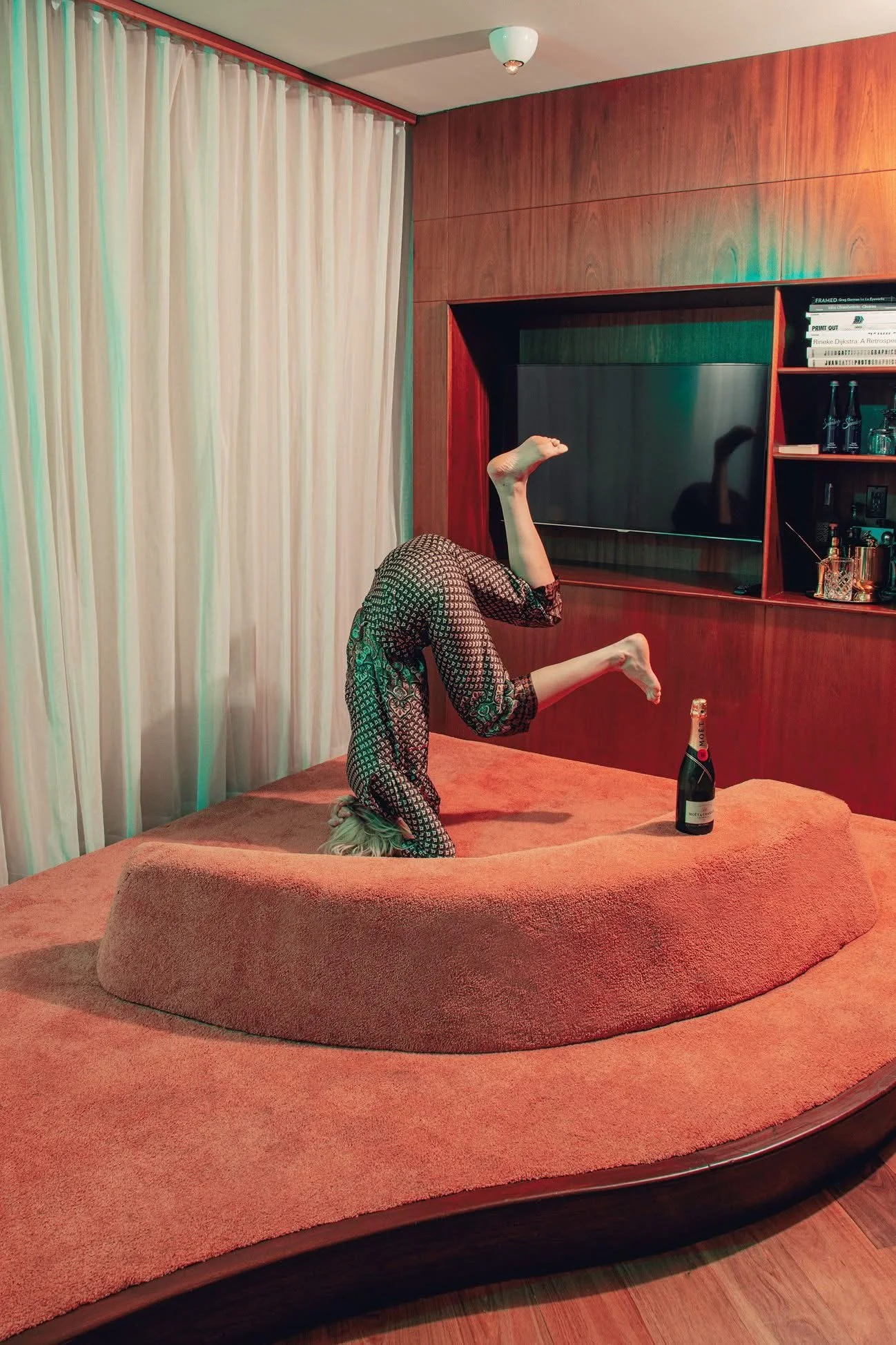A person wearing patterned pajamas is balancing on their head on an orange carpeted bed, with one leg raised and the other bent, holding a bottle of Moët & Chandon champagne. The room has a wooden wall with a black TV, shelves with books and bottles, and cream curtains.