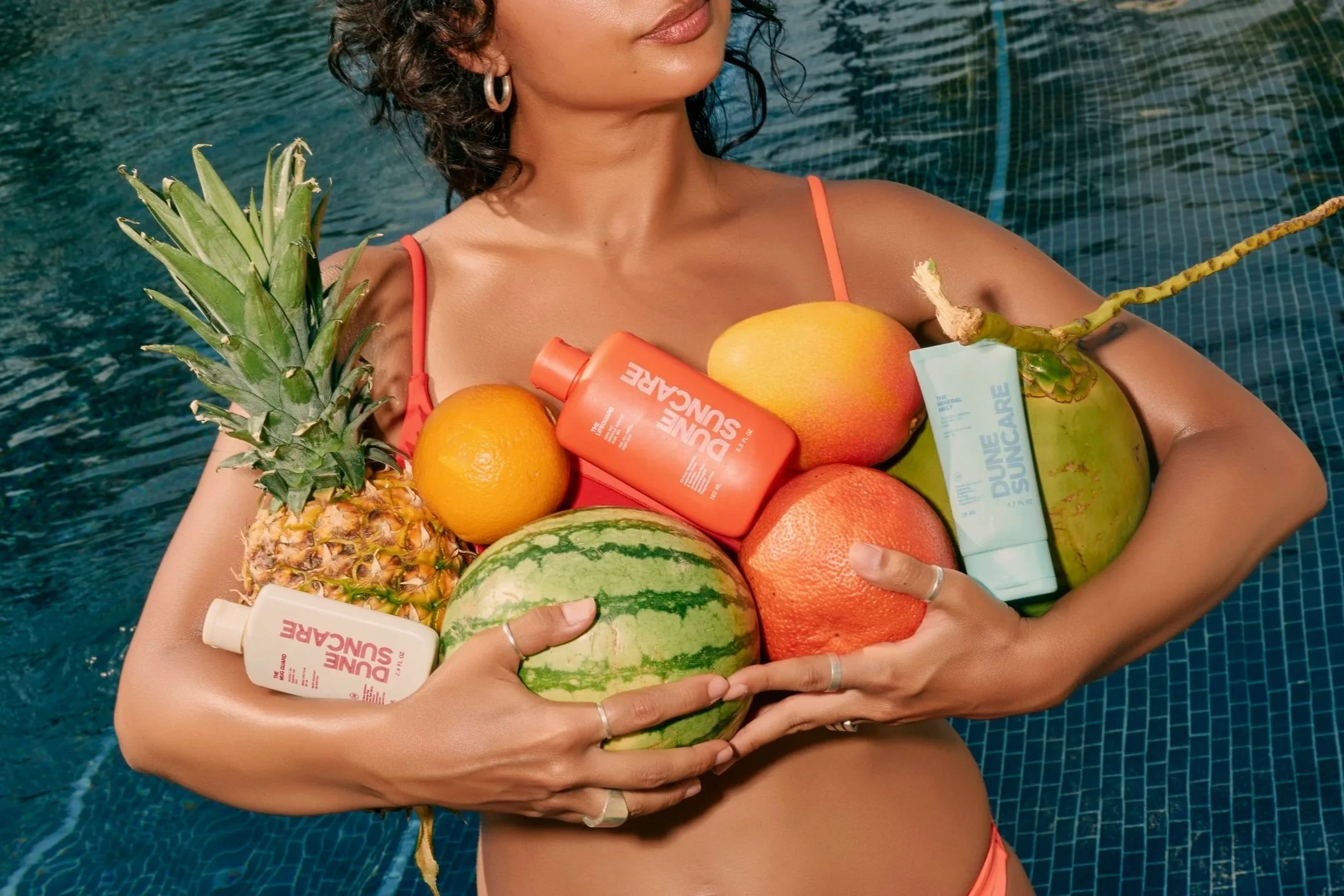 Woman in a swimsuit holding various fruits and skincare products by a pool.