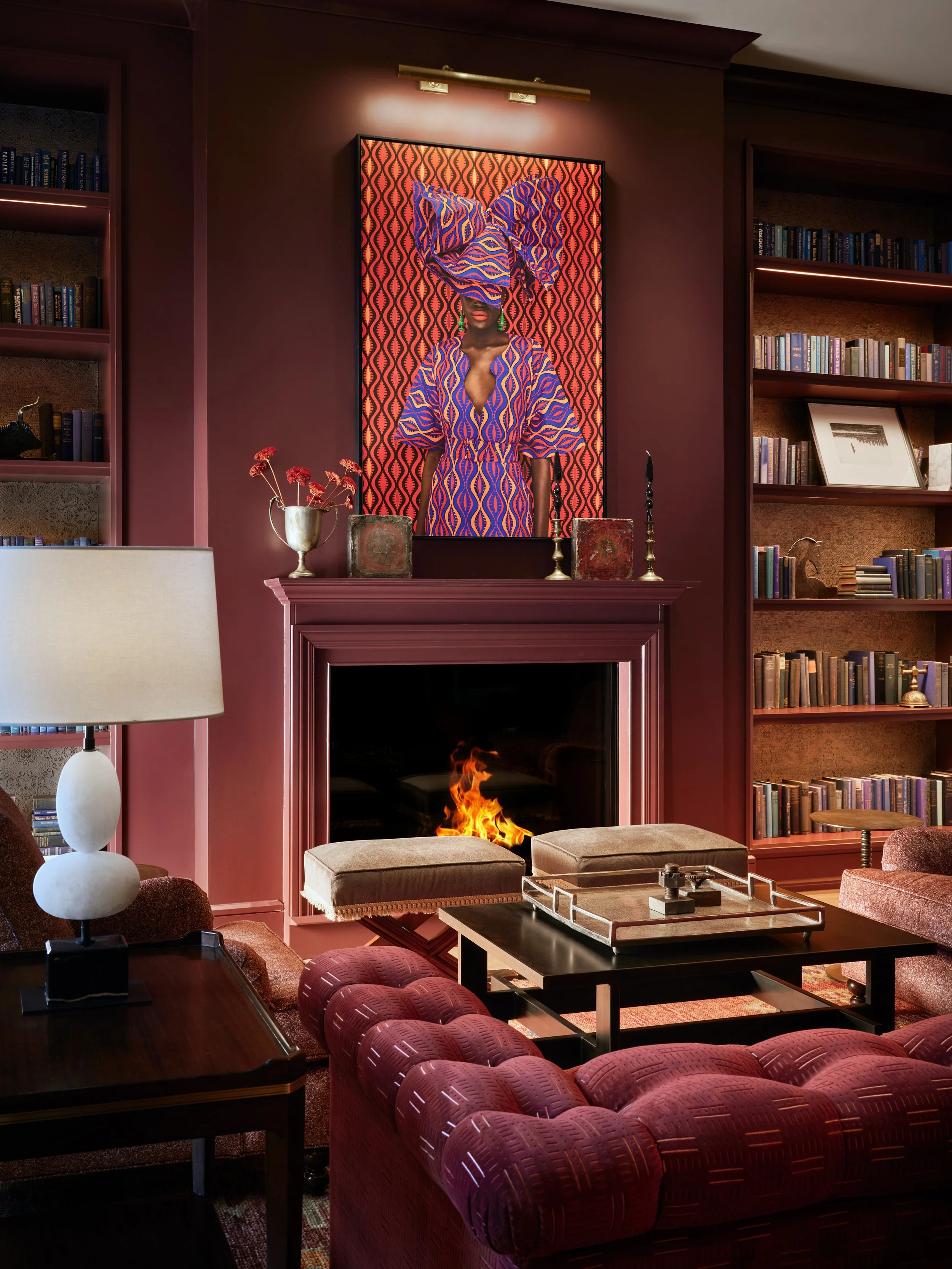 A cozy living room with a pink fireplace, a fire burning, surrounded by a large red upholstered armchair and a sofa. There are bookshelves filled with books, a table with a tray, candles, and framed artwork. A large painting of a woman with a headwrap and dress in patterned fabric decorates the wall above the fireplace, with a lamp above it illuminating the painting.