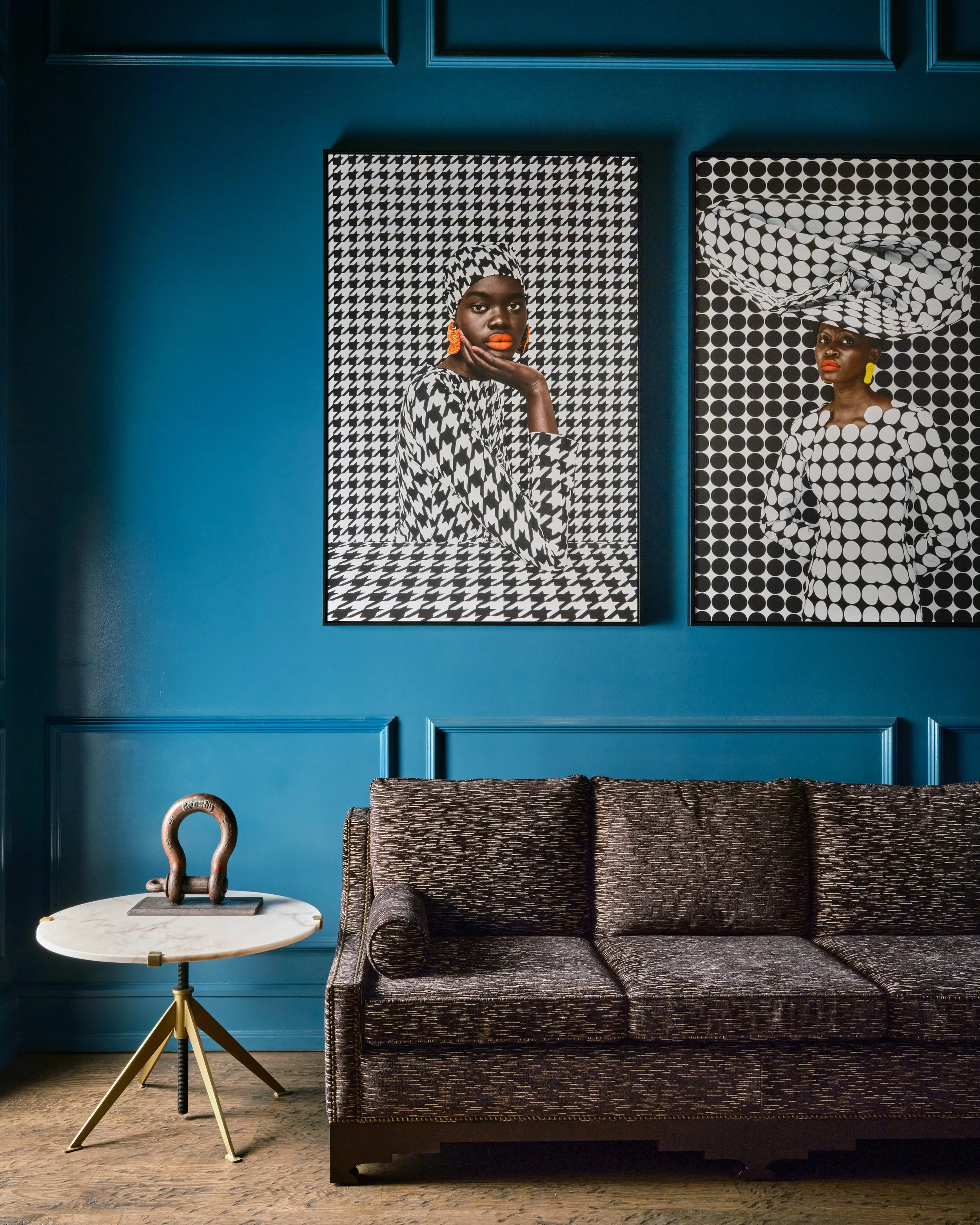 Interior of a room with a blue wall, decorated with two large black-and-white patterned portraits of women with bold red lipstick and earrings. Below the portraits is a dark textured sofa, and to the left, a small round marble-top side table with a brass and wood sculpture on it.