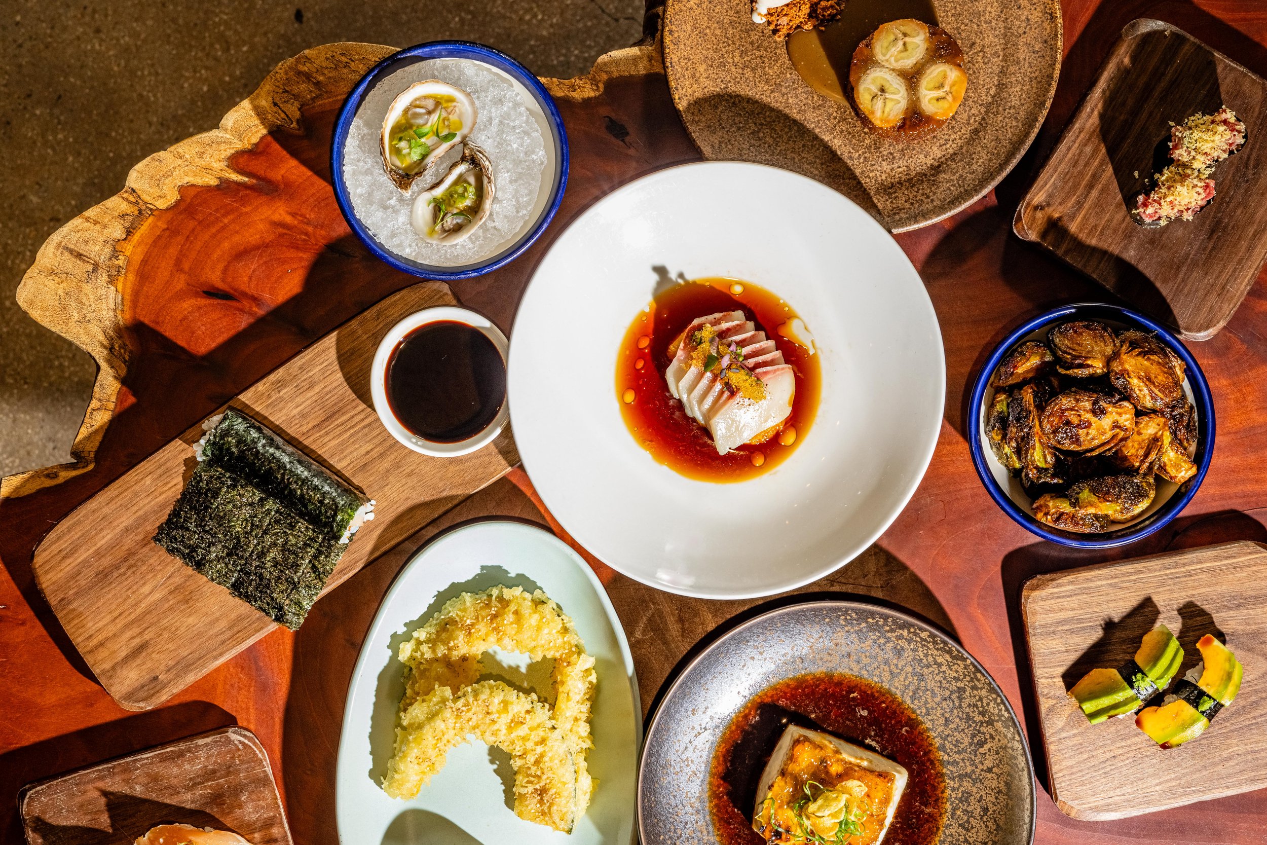 A variety of Asian dishes on a wooden table, including sushi rolls, sashimi, oysters, grilled seafood, tempura, and dipping sauces.