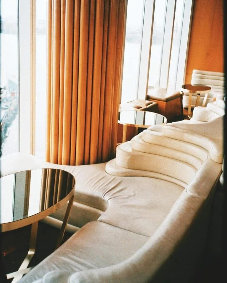 Elegant lounge with cream-colored sofa, round mirrored tables, large floor-to-ceiling windows, and wooden accents.