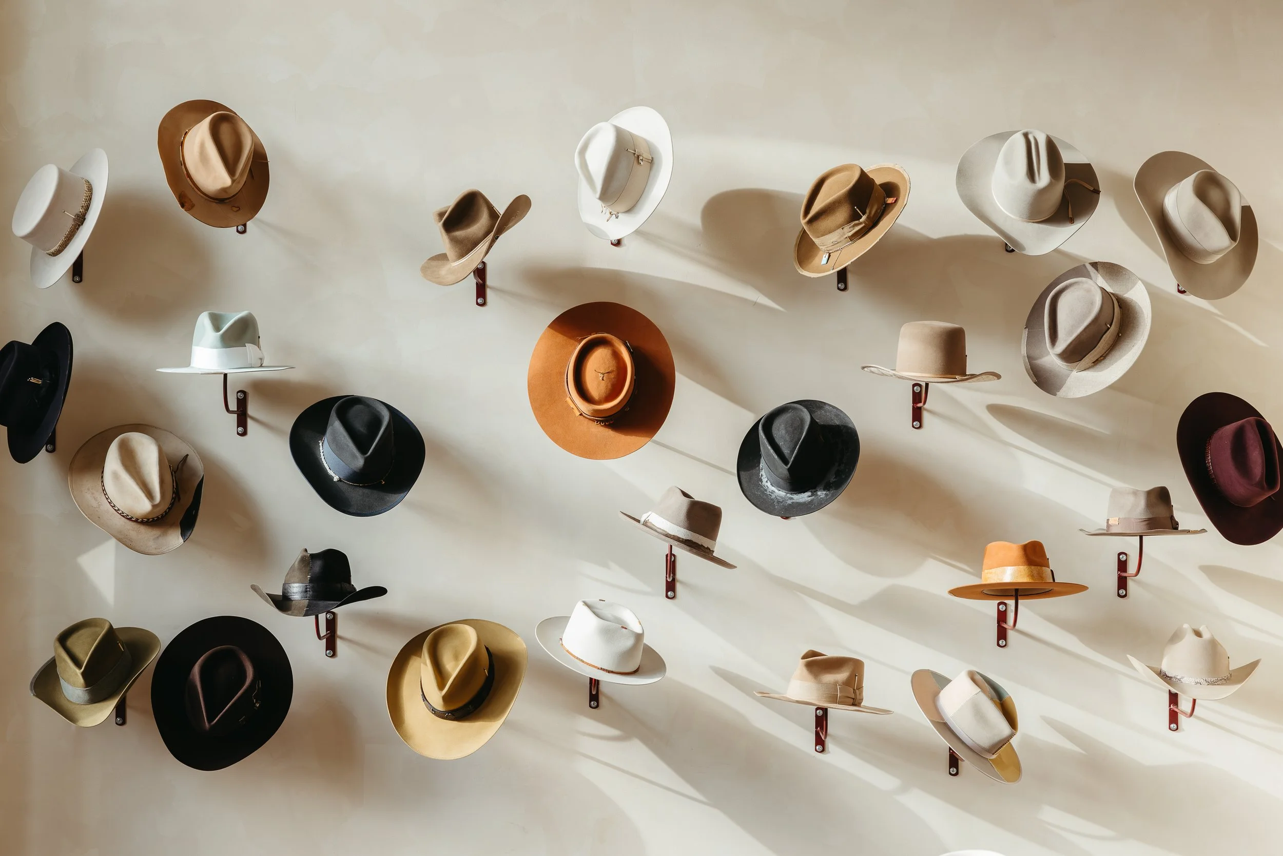 A display of various cowboy hats mounted on a white wall, arranged on metal hooks in different colors and styles.