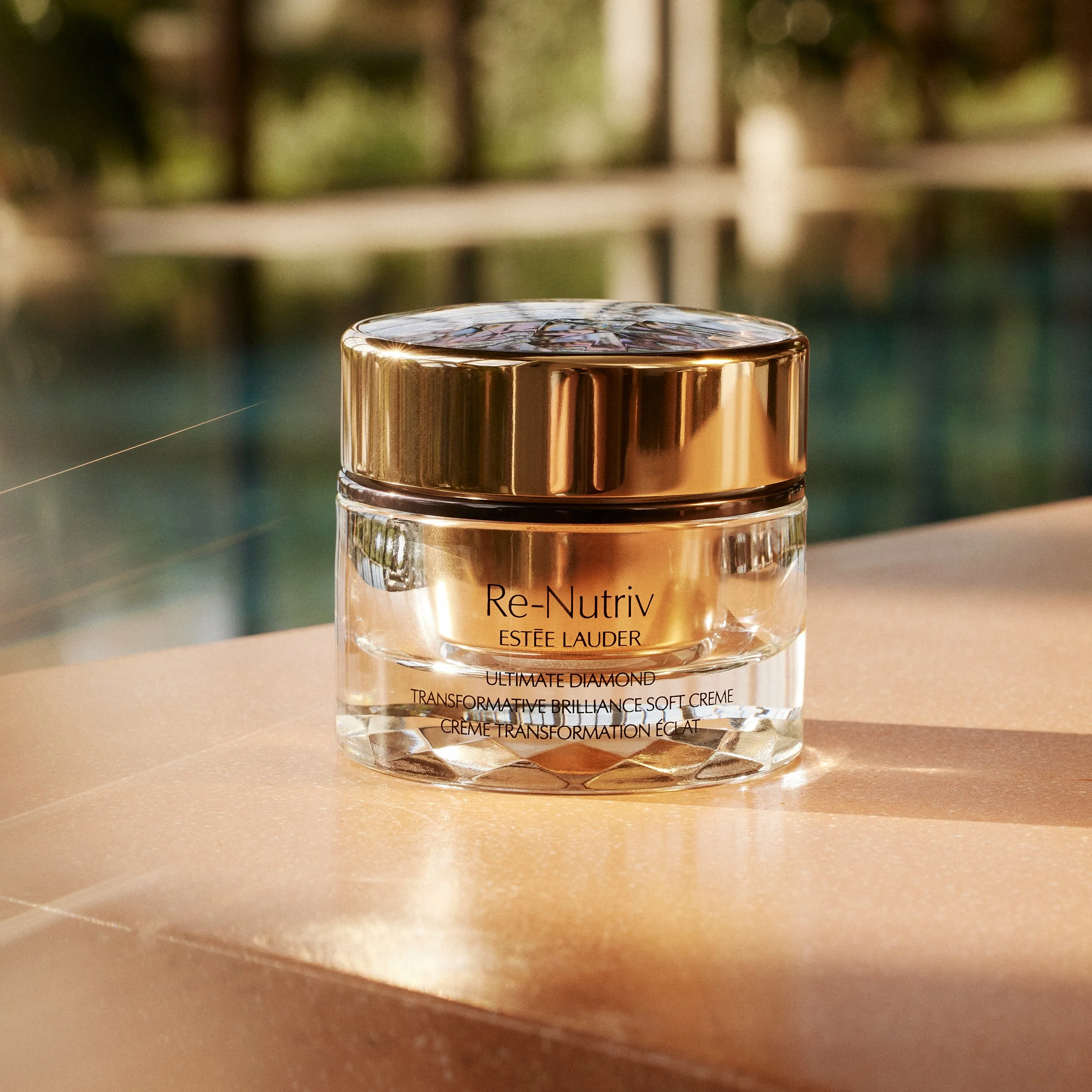 Close-up of a jar of Estée Lauder Re-Nutriv Ultimate Diamond Transformative Brilliance Soft Creme with a reflective, gold-colored lid, on a beige surface with a blurred background.