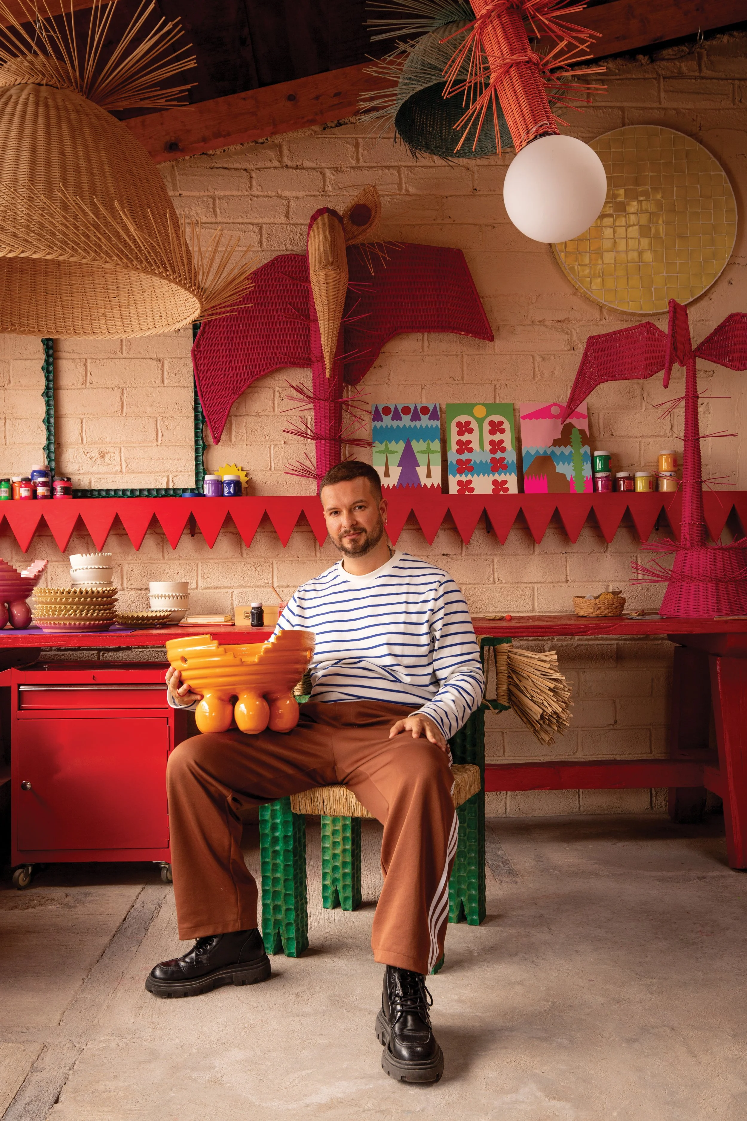 A man with a beard and mustache sitting in a colorful room, holding a large orange ceramic bowl with a face design. He is wearing a white and blue striped shirt, brown pants, and black boots. The room's background features bright decorations, including abstract paintings, large woven lampshades, and tropical-themed decor, with a red counter and various small jars and containers.