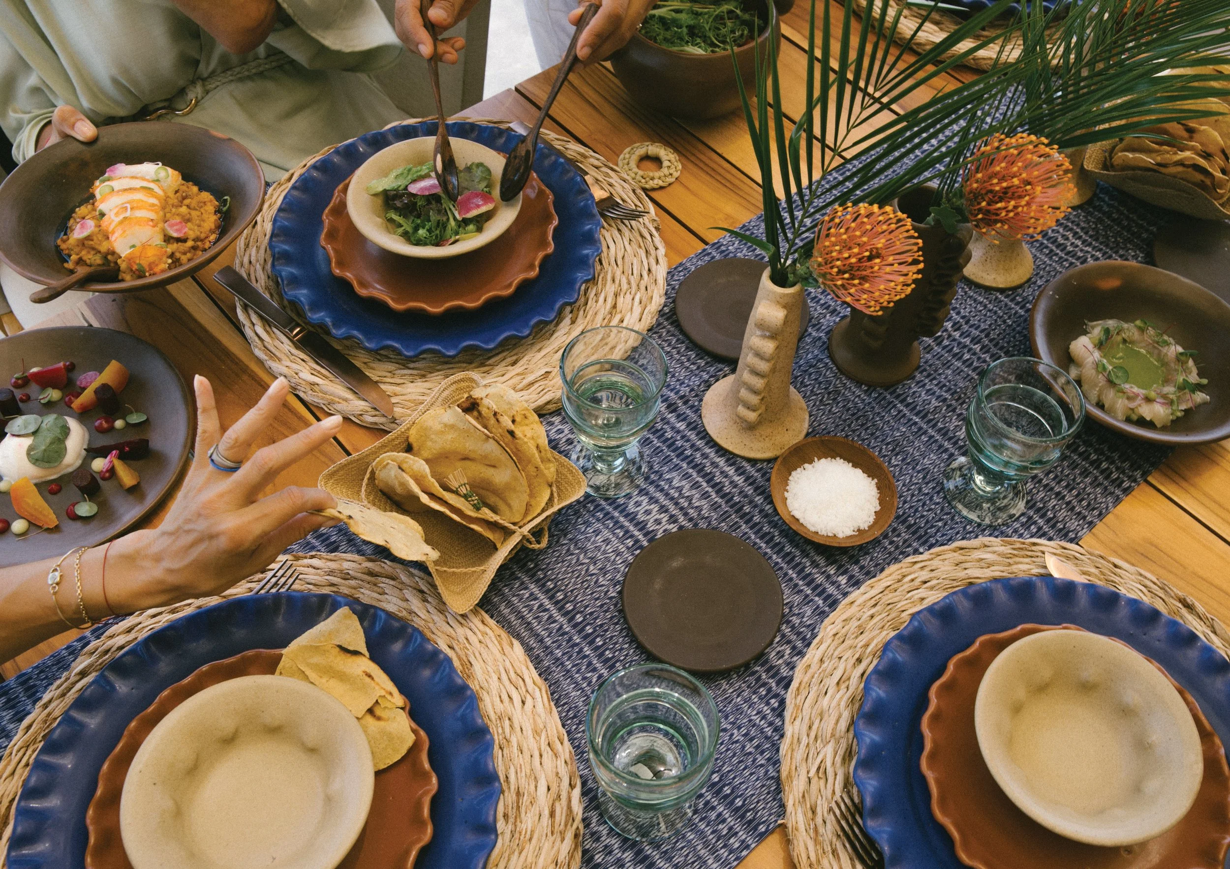 A table set with various dishes including bowls of soup and salads, decorated with vases of flowers, with hands visible reaching for food, and glasses of water on a blue tablecloth.