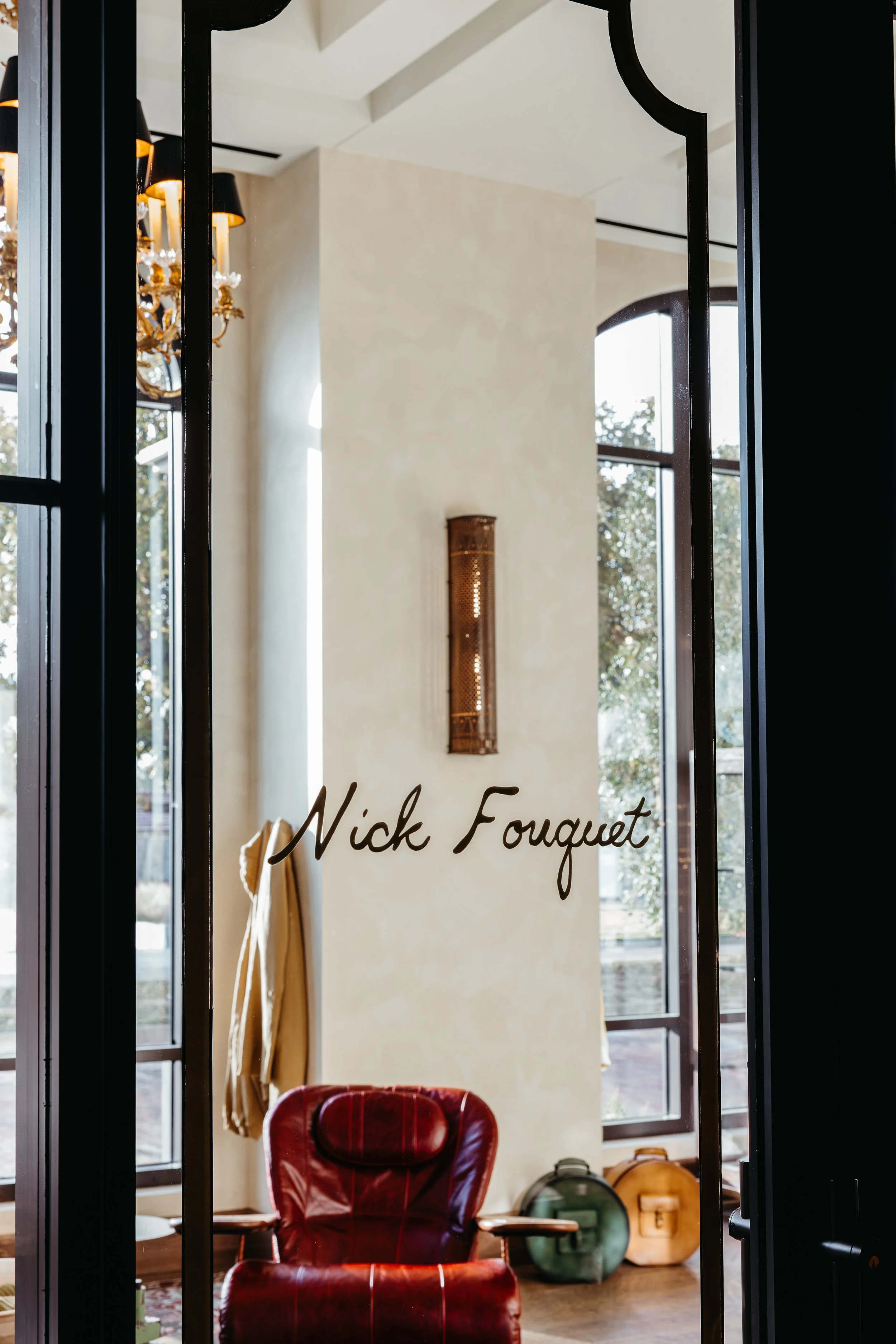 View through glass door with the words 'Nick Fouquet' written on it, showing a cozy interior with a red leather armchair, a beige coat hanging on a hook, decorative bags and lanterns near a large window, and a chandelier hanging from the ceiling.