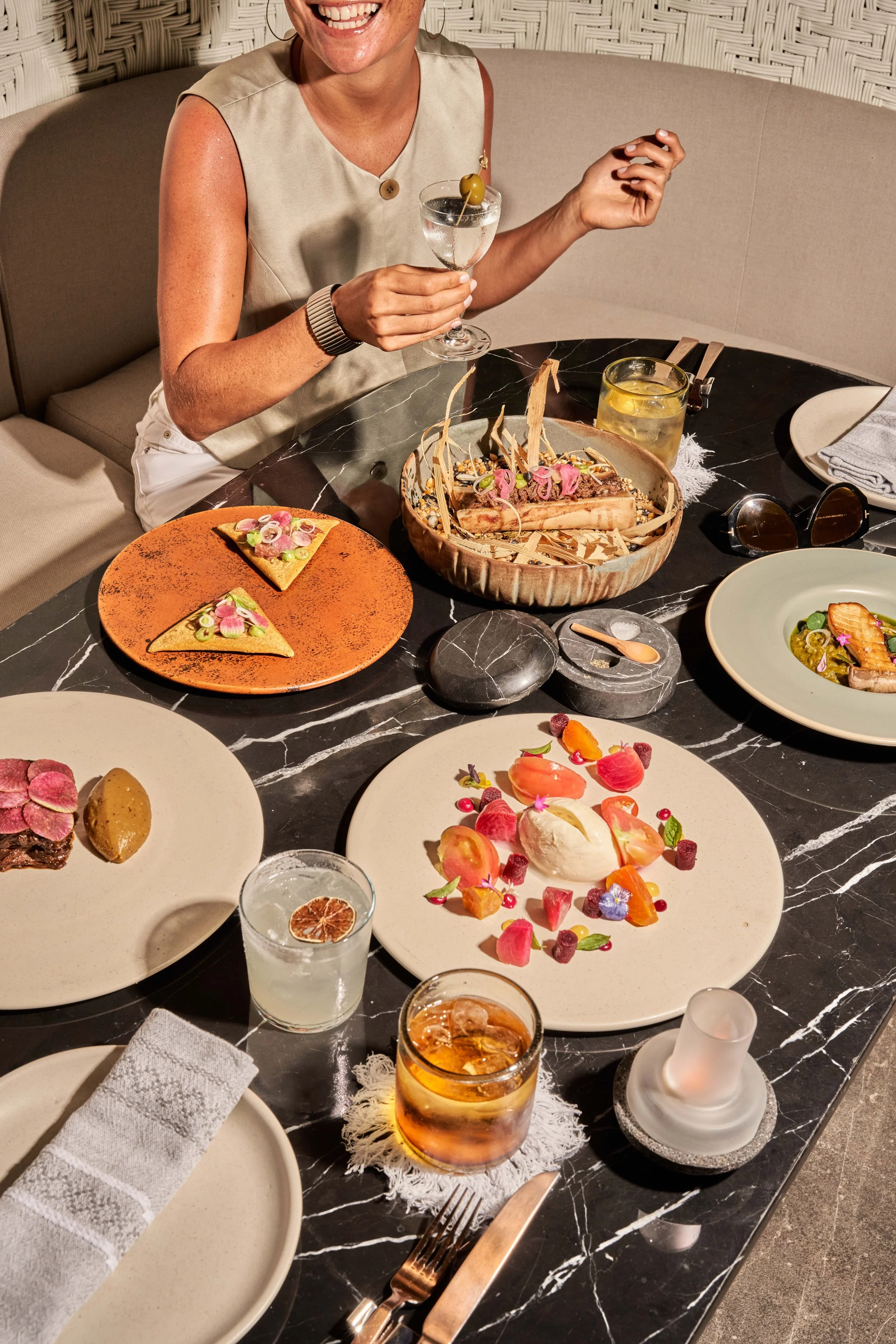 A woman smiling at a dinner table with various gourmet dishes, glasses of water and whiskey, and decorative plating of desserts.