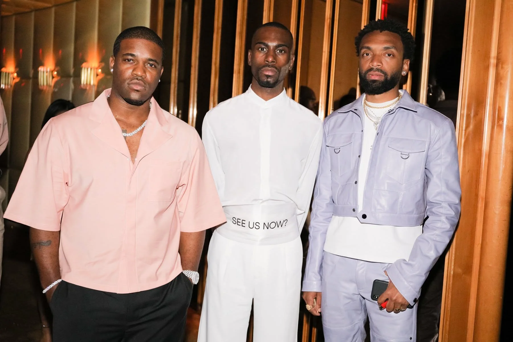 Three men standing in a row at an indoor event, dressed in stylish outfits with accessories, against a wooden wall background.
