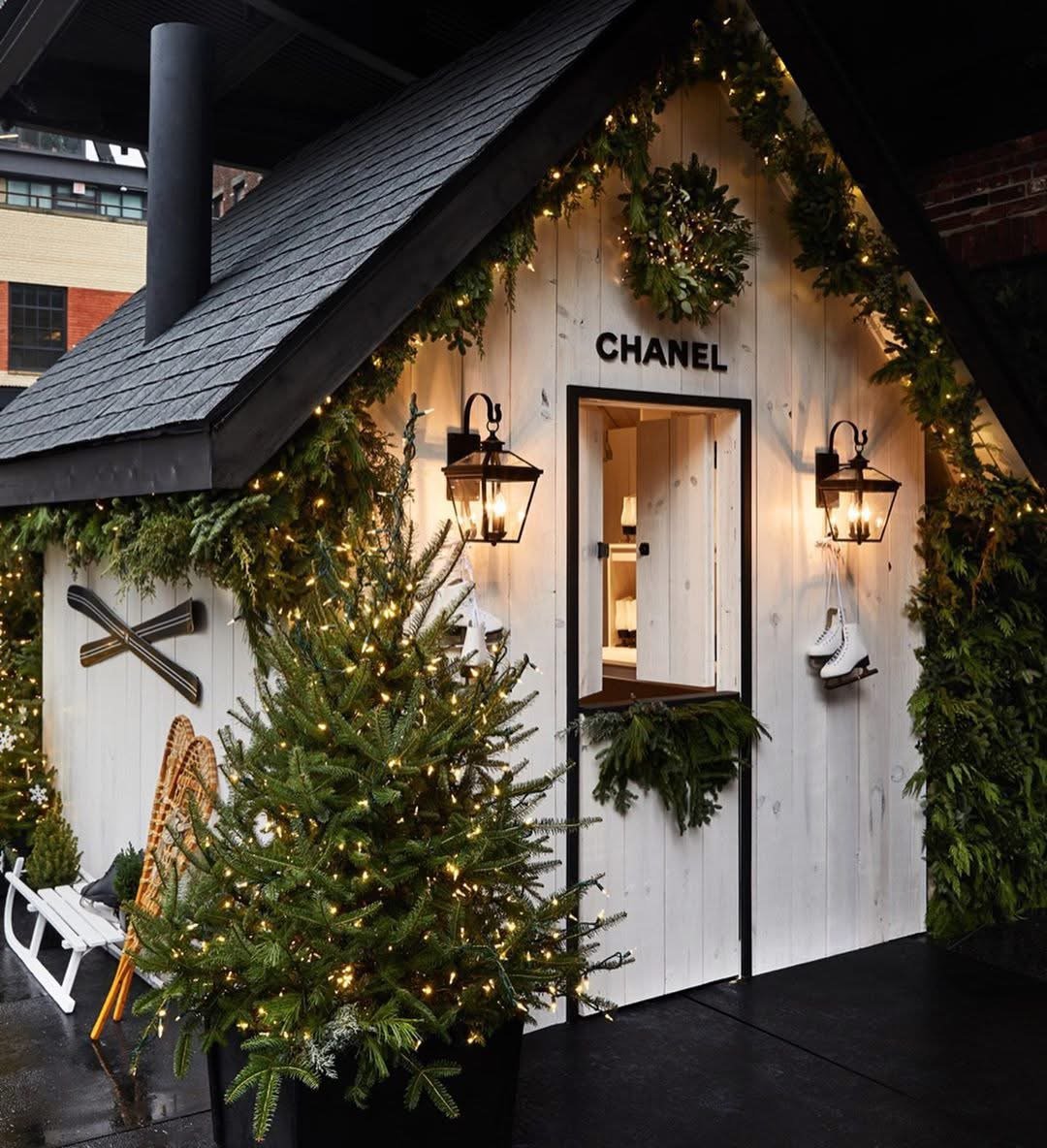 Christmas-themed outdoor chalet with a 'CHANEL' sign, decorated with wreaths, garlands, and string lights, featuring a small Christmas tree with lights, ice skates hanging on the wall, and vintage lanterns.