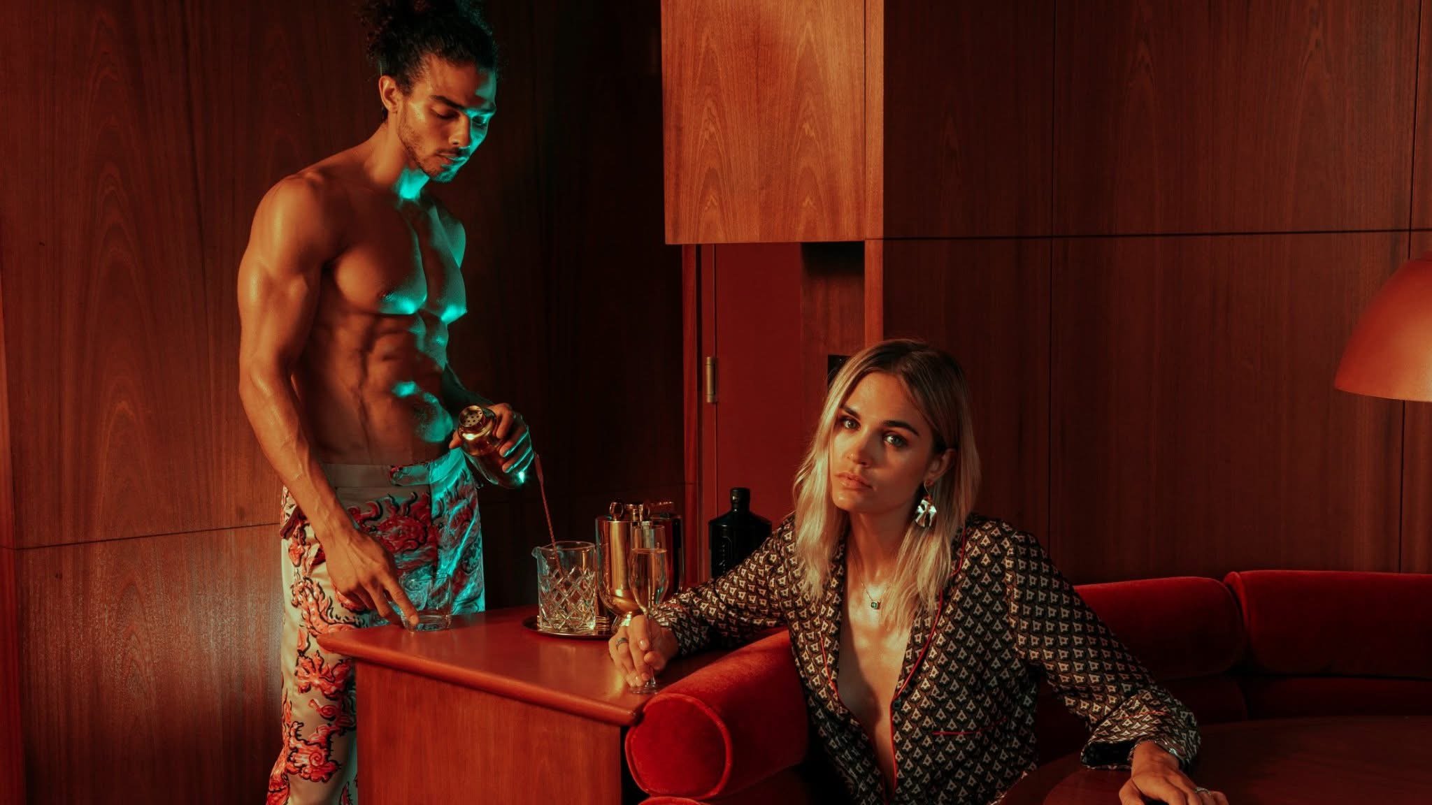 A shirtless man pouring a drink at a bar or restaurant while a woman with long blonde hair and a patterned blouse sits at the table, looking at the camera. The background features wooden paneling and a red lamp.