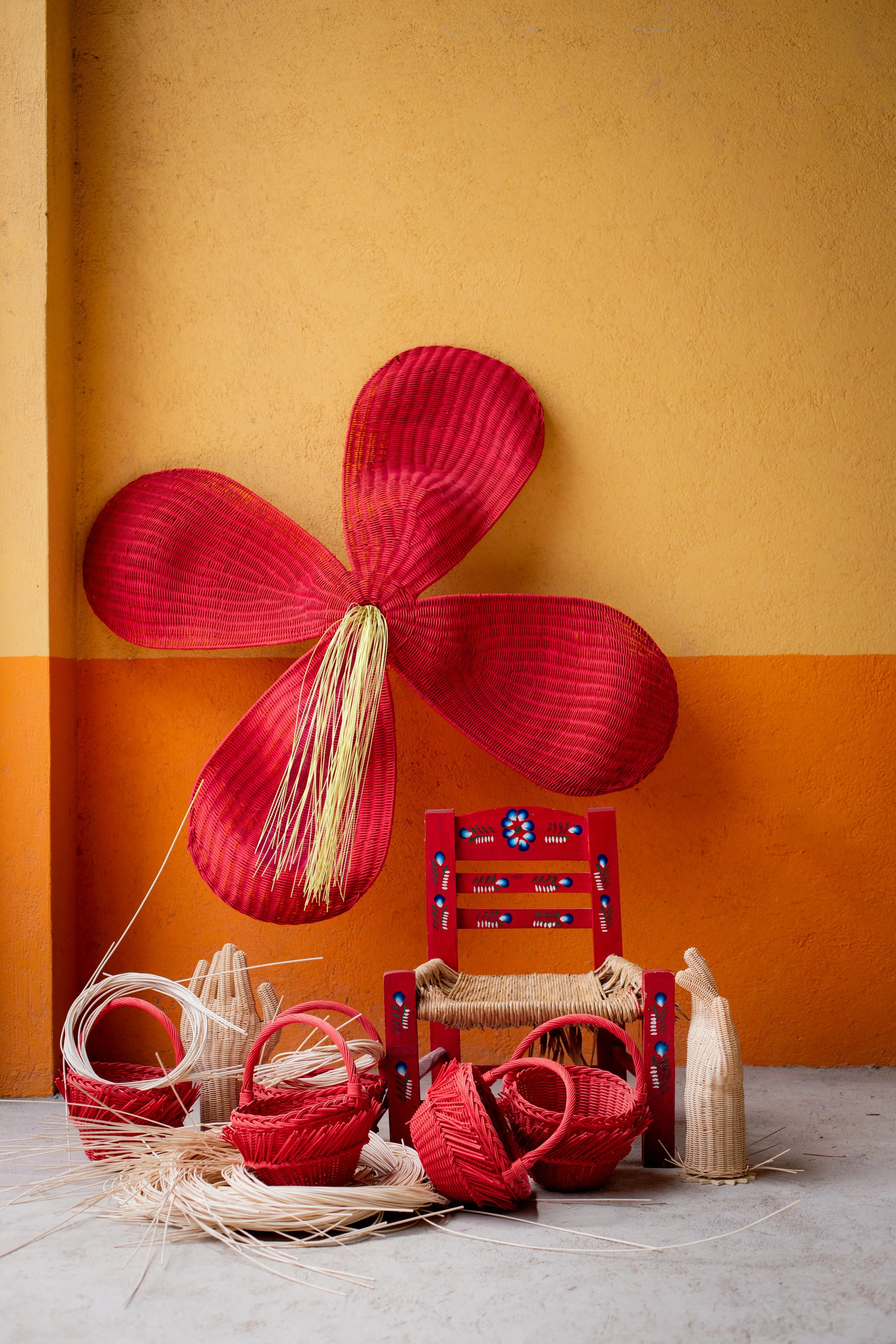 Display of red woven baskets, straw hats, and a red and natural-colored woven chair against an orange wall.