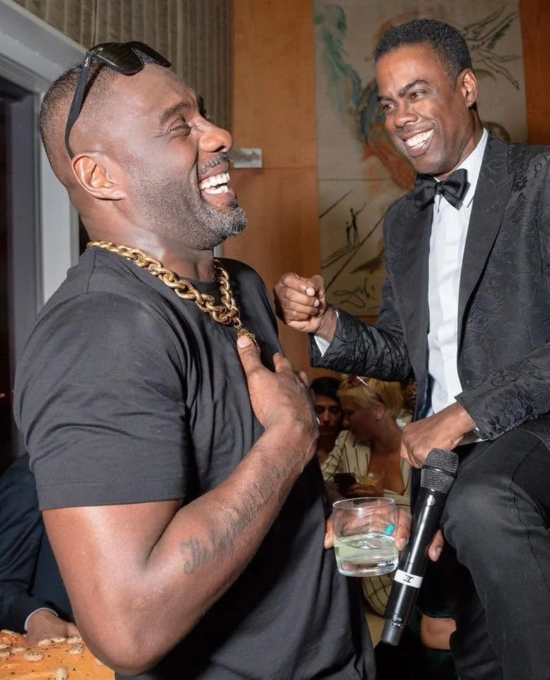 Two men are laughing and smiling at each other during a social event. One man is wearing a black T-shirt with sunglasses on his head, a gold chain, and has tattoos on his arm. The other man is in a tuxedo with a bow tie, holding a microphone and a glass of water. They seem to be enjoying a fun moment together.