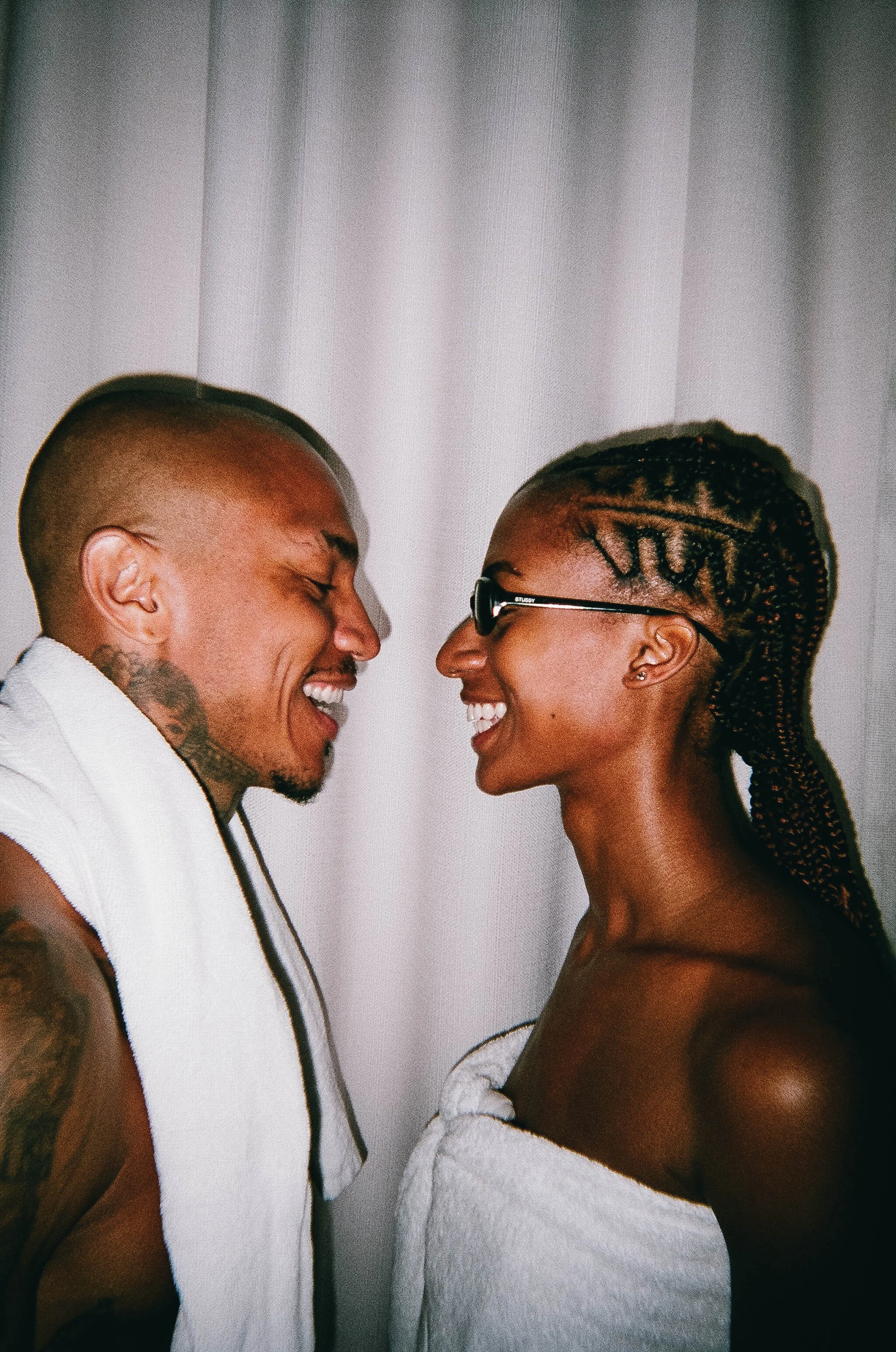 A man and woman facing each other, smiling and laughing, wrapped in white towels, with a plain white curtain in the background.