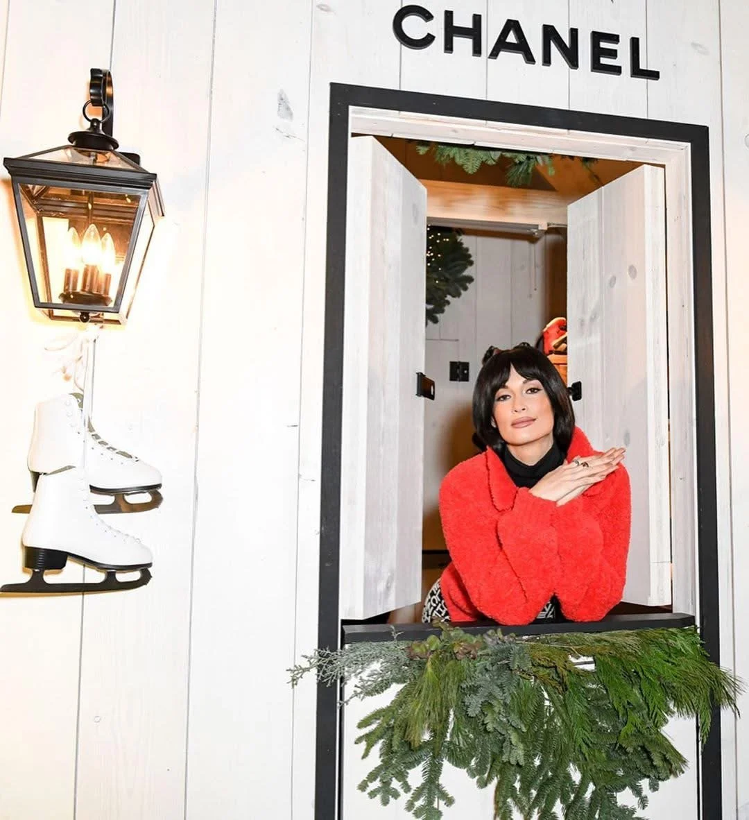 Woman with short black hair, wearing a red sweater, leaning on a window frame decorated with green pine branches, inside a store with a "CHANEL" sign above. Decor includes a wall-mounted lantern and ice skates on the wall.
