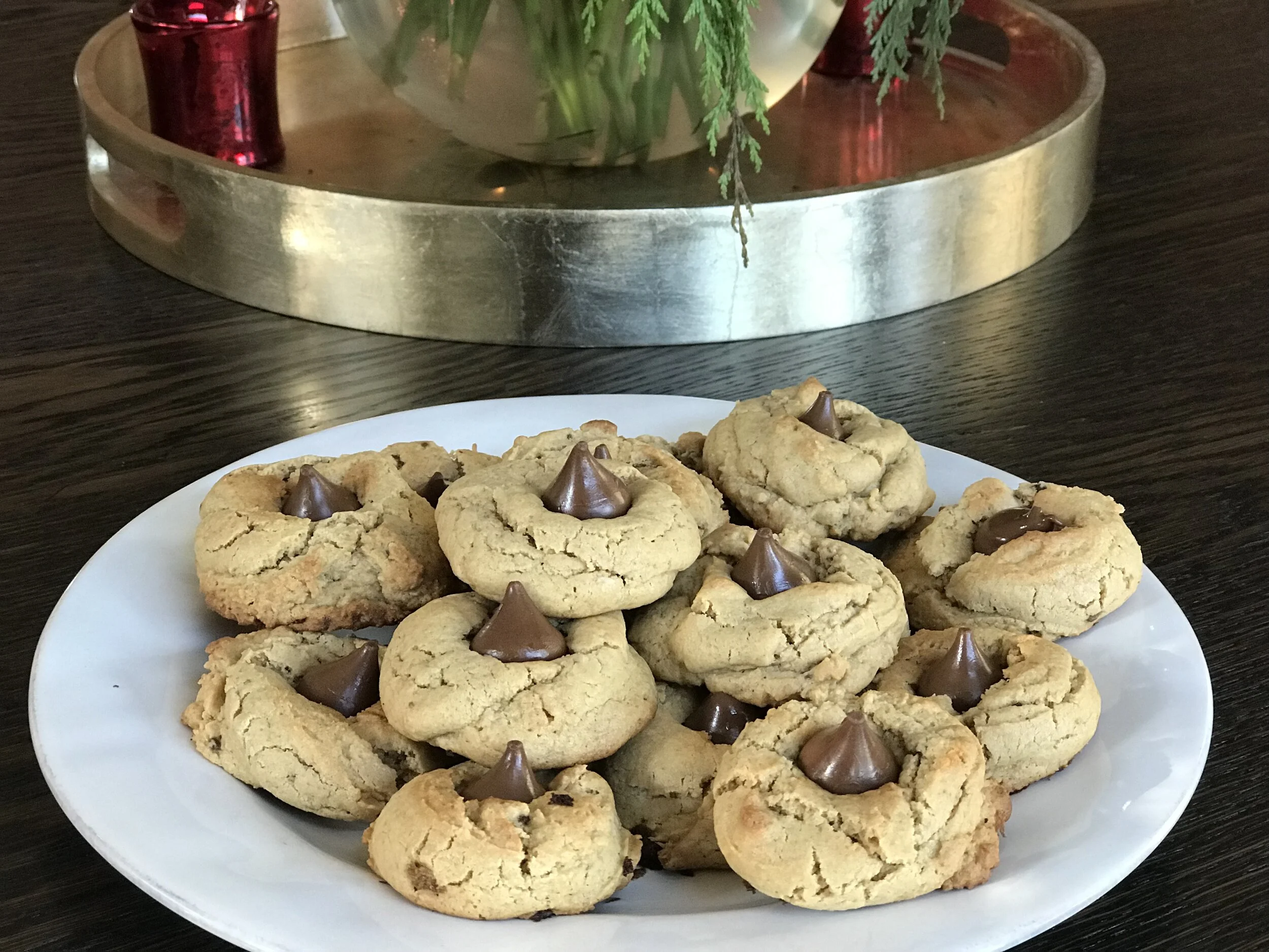 Plate of chocolate chip cookies with chocolate kisses on a dark wooden table, with a decorative tray with foliage in the background.