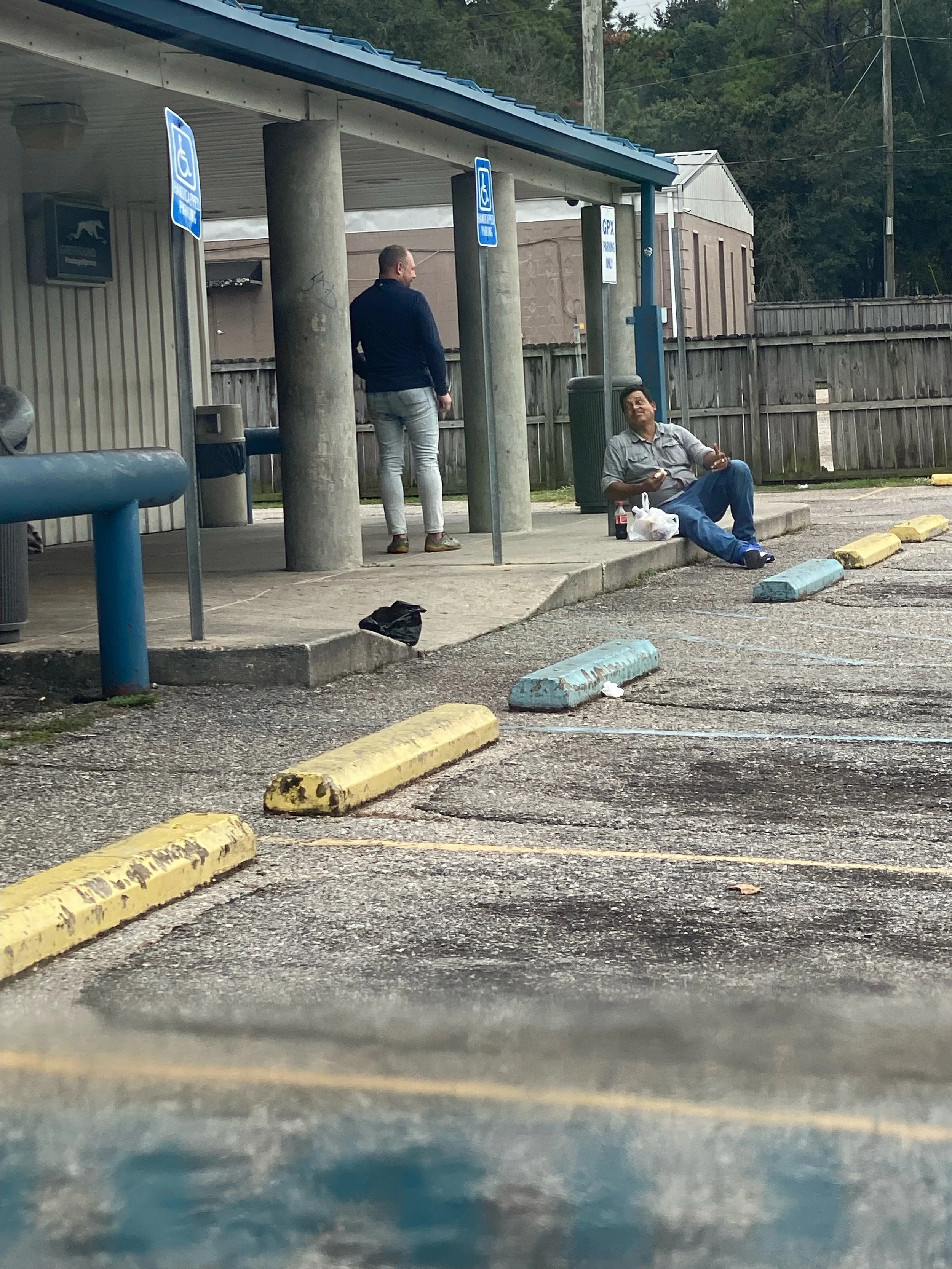 Two men pray in a parking lot outside a building, one sitting on the curb eating, the other standing near the building.