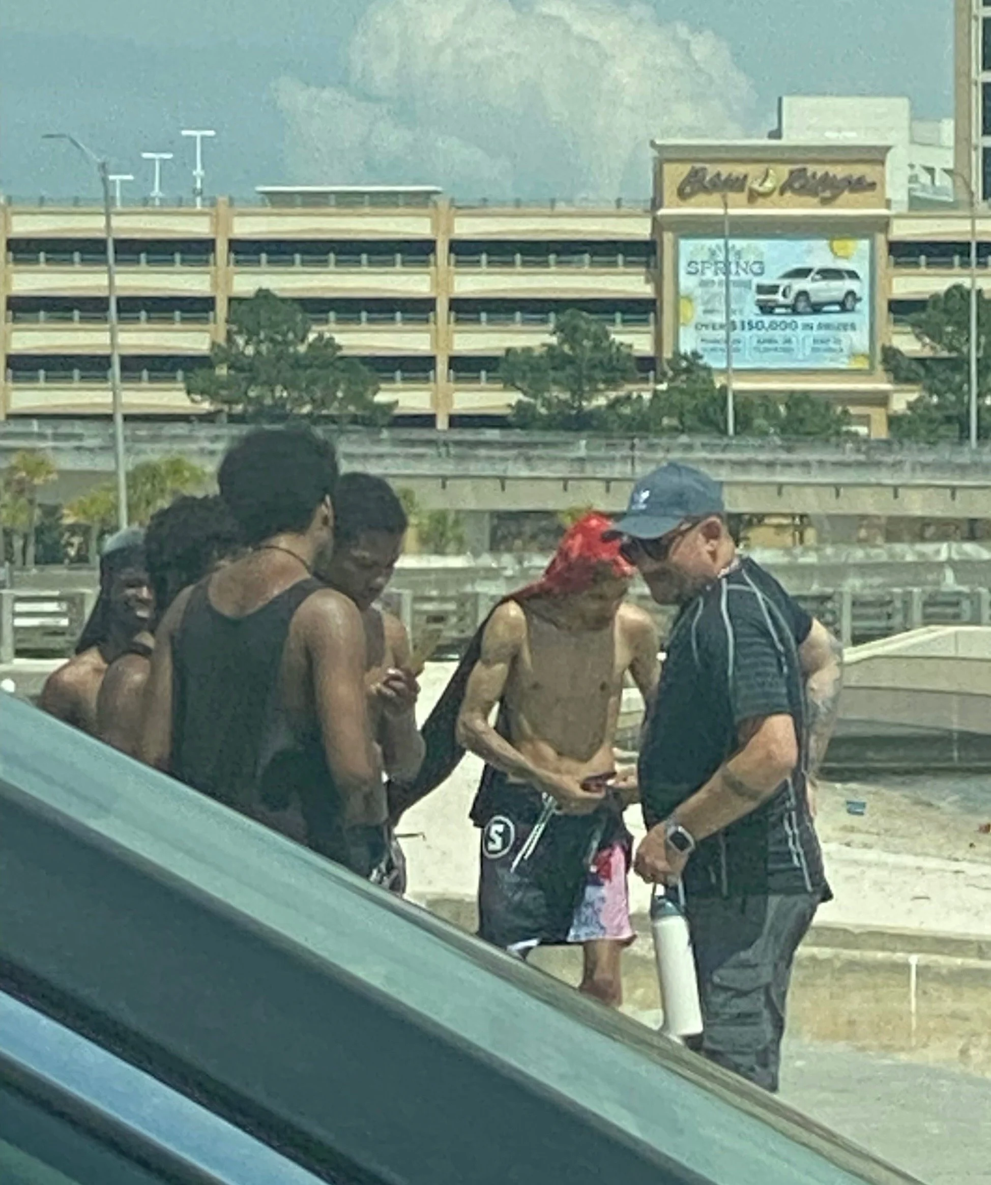 Group of people standing outdoors near a parking lot, some shirtless, with one man wearing sunglasses and a cap, holding a water bottle.