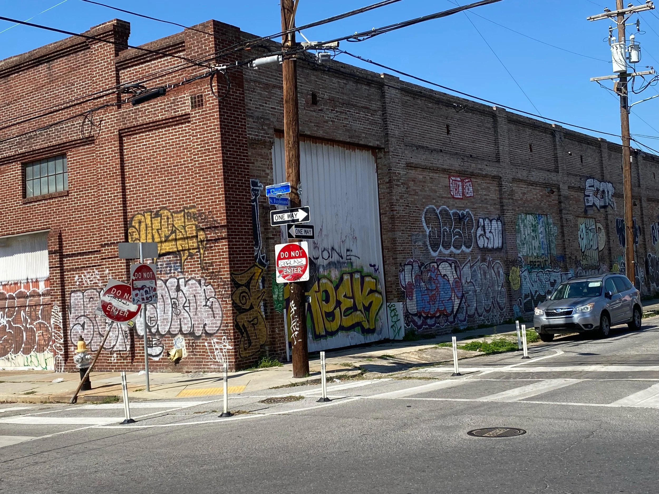 Street corner with graffiti-covered brick wall, multiple stop and do not enter signs, parked gray car, utility poles, and overhead power lines under a clear blue sky.