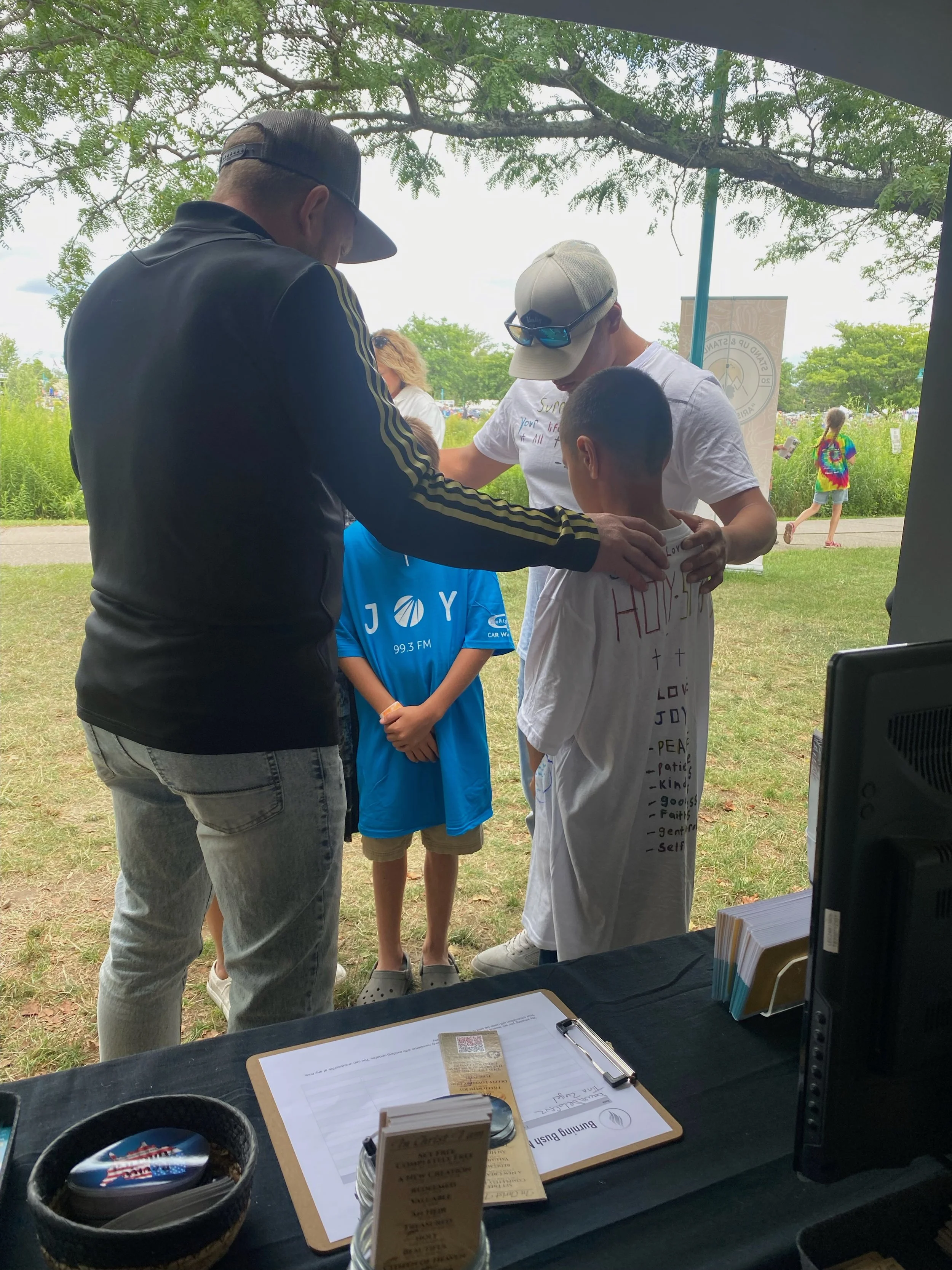A group of people, including a man and several children, are gathered outdoors under a tree during daytime. The man appears to be giving something to one of the children, who is dressed in a white shirt with handwritten words. A woman and other child