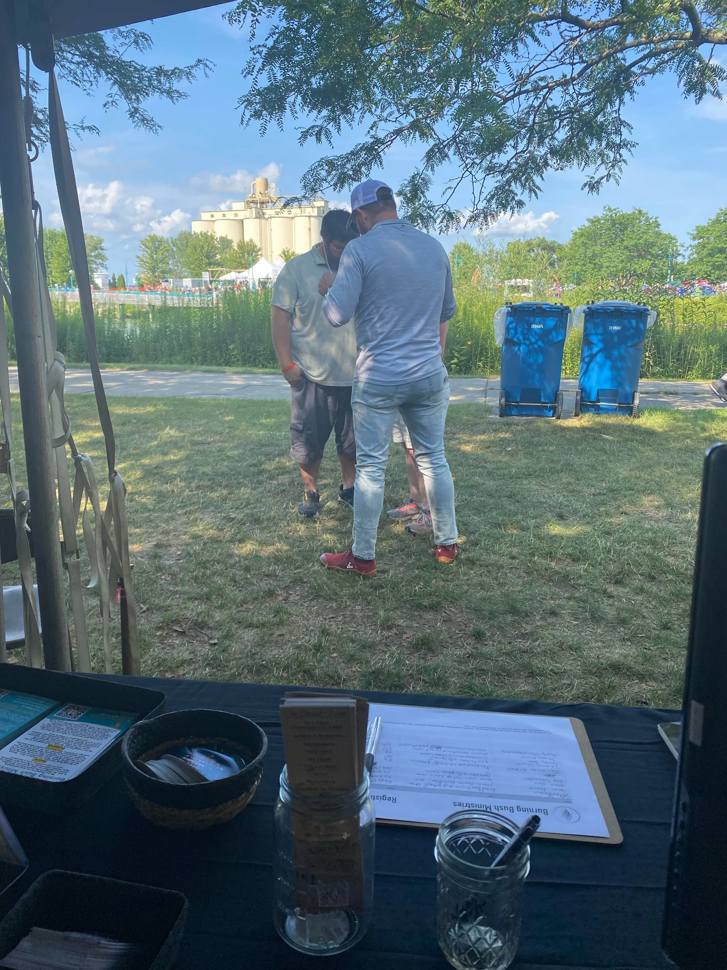 People gathered outdoors under a tree, engaged in conversation near two blue trash bins, with a grassy area and buildings in the background. The photo is taken from inside a tent or booth facing the scene, with papers, a jar, and other items on a tab