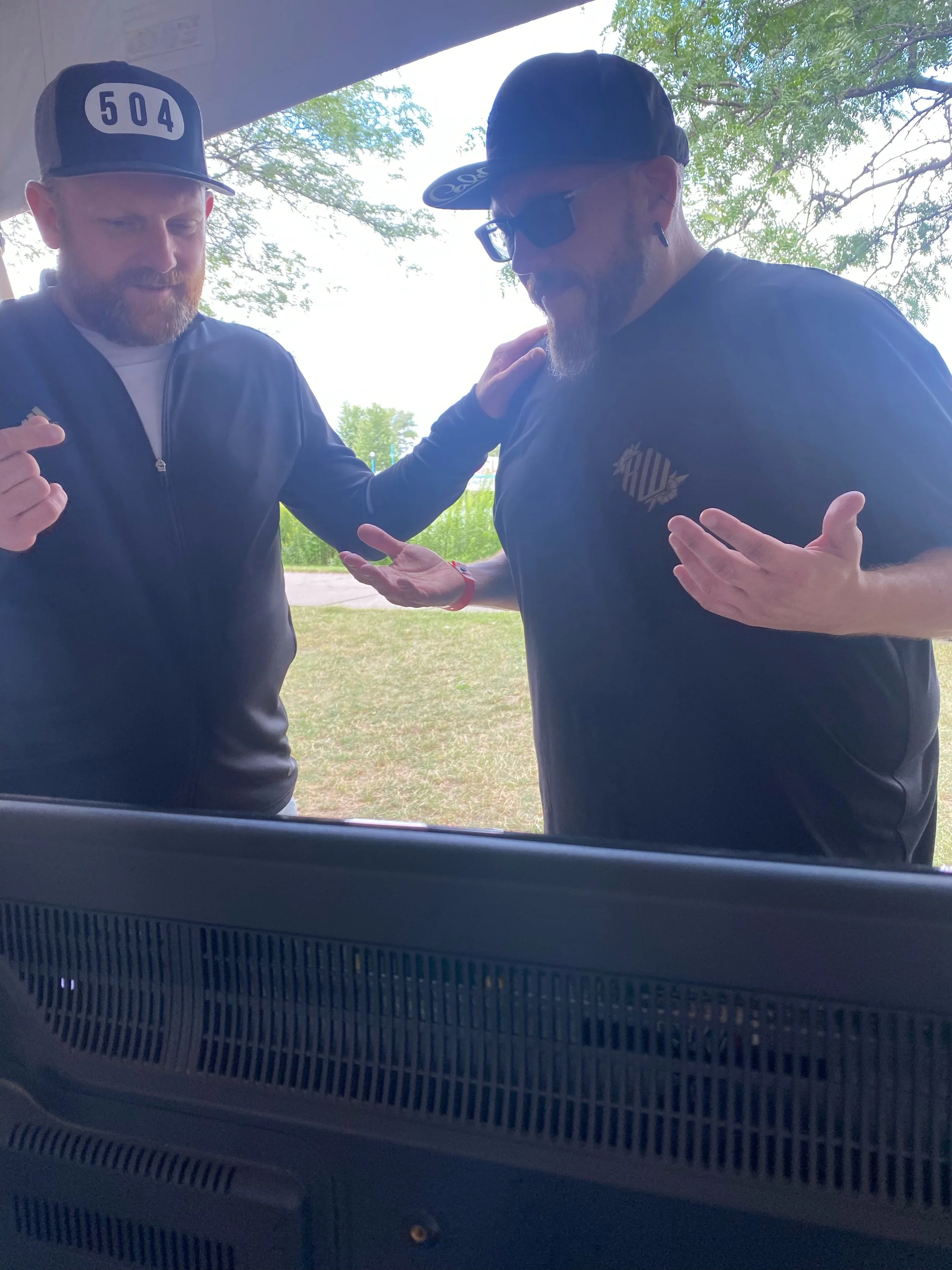 Two men standing outside under a shelter, engaging in a conversation with one man resting his hand on the other's shoulder. One man is wearing a black baseball cap with '504' on it, a black jacket, and has a beard. The other man is wearing a black ca