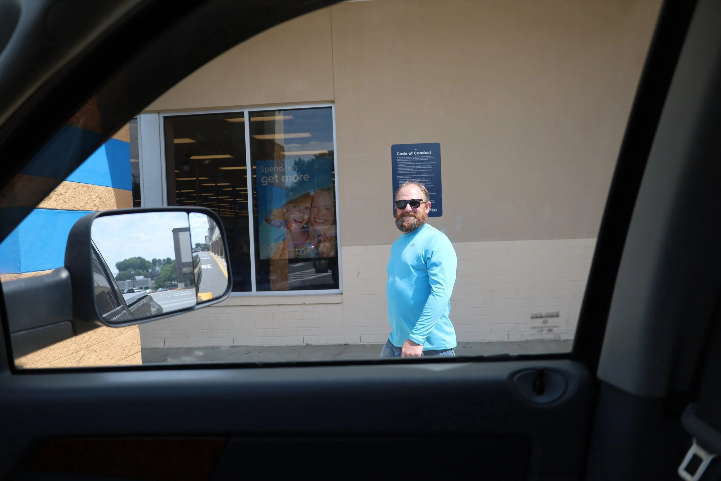 Man with beard and sunglasses in blue shirt walking outside a store, seen through car window and side mirror.