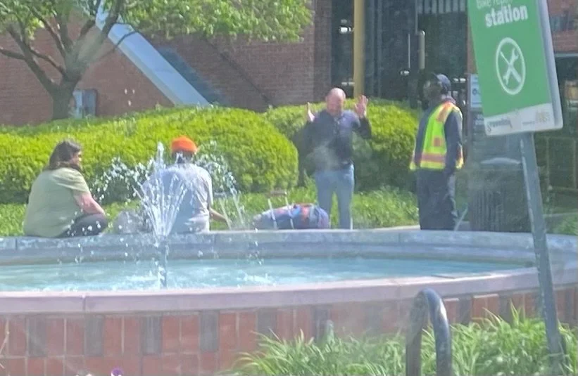 A group of four people gathered near a fountain outside, with one person in a yellow reflective vest and a person standing with hands raised, possibly talking or explaining something. A sign indicates a train station nearby.
