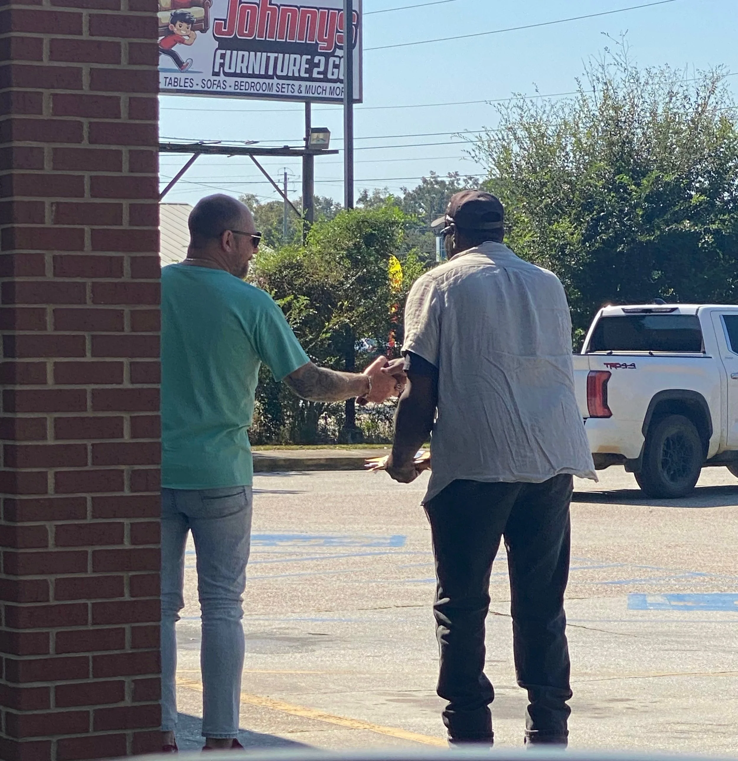 Two men are standing outside in a parking lot, with a white pickup truck in the background. One is wearing a light-colored short-sleeve shirt and a hat, and the other is wearing a turquoise shirt with tattoos on his arm. A brick wall is partially vis