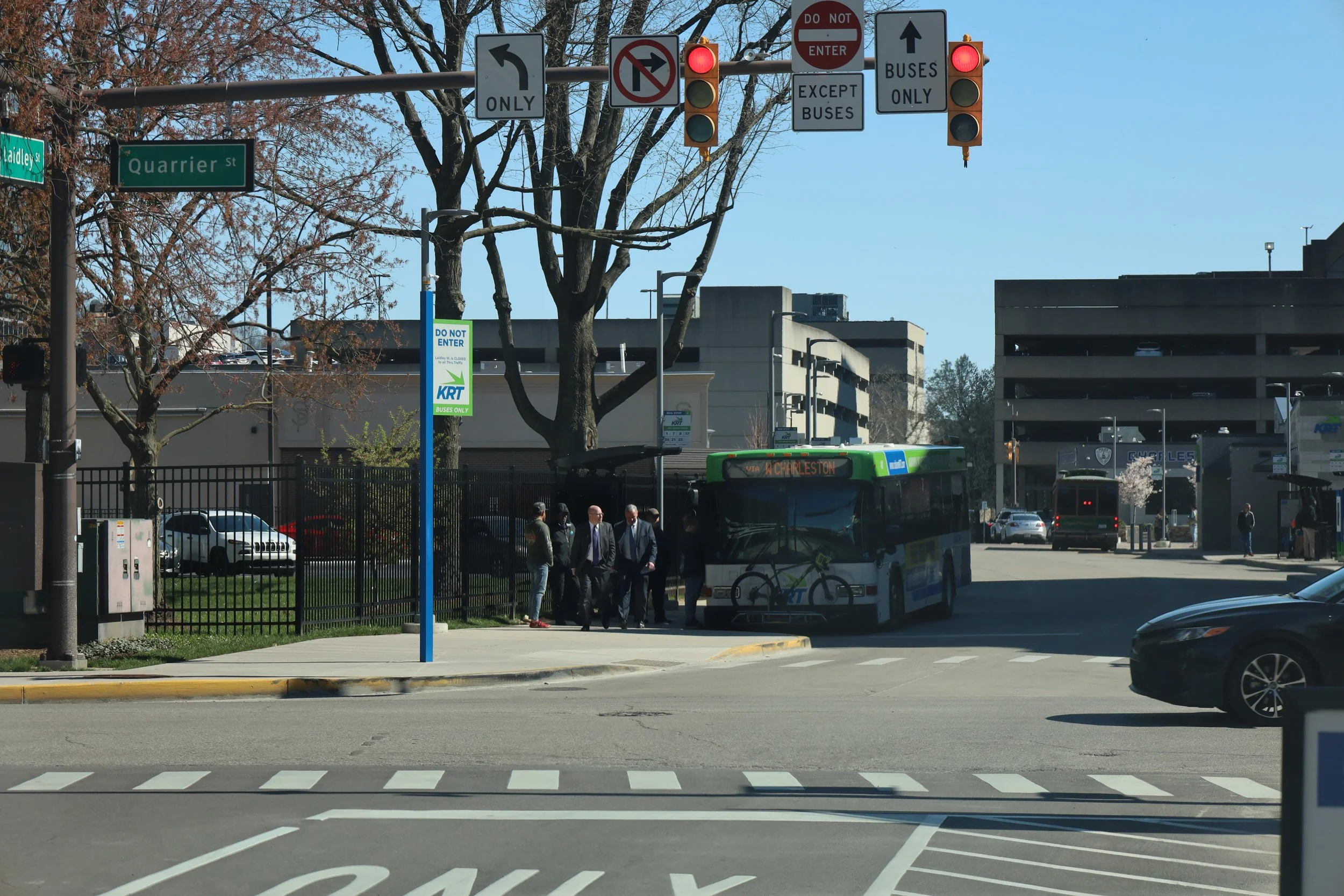 Street scene with traffic lights, signs, a bus labeled 'Charleston', a group of people waiting at a bus stop, trees, parked cars, and buildings in the background.